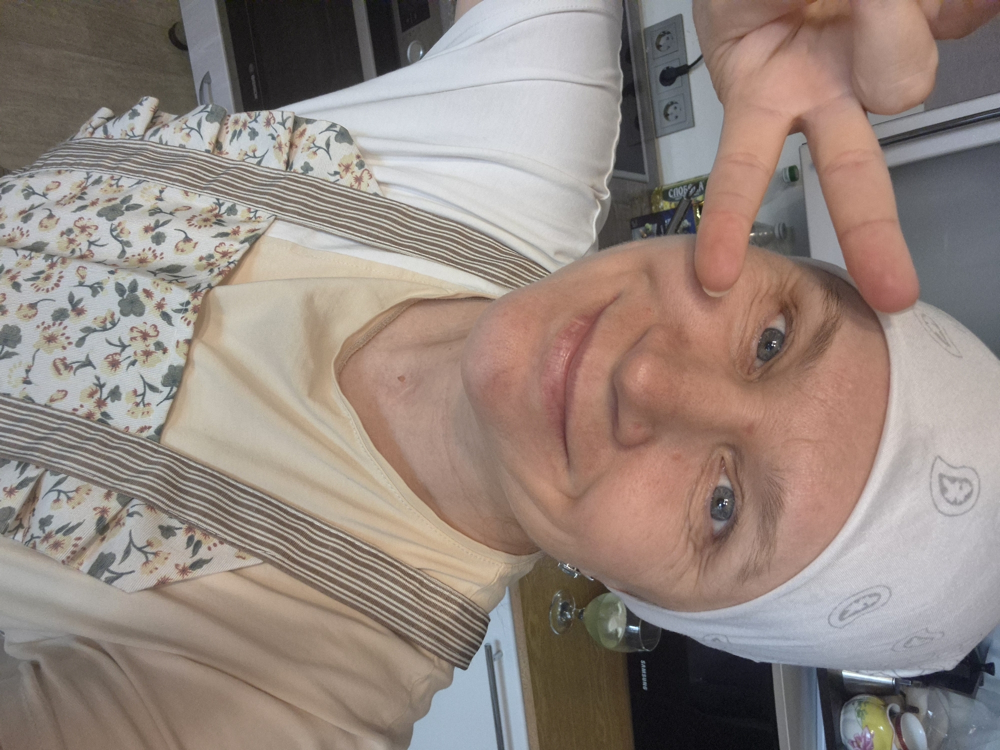 A woman is taking a selfie in a kitchen. She is wearing a white bandana, a light-colored t-shirt, and a floral apron. She is holding up two fingers in a peace sign with her right hand. In the background, there is a refrigerator, a countertop with kitchen appliances, and a glass of drink. The woman appears to be smiling and looking at the camera. The image does not provide enough information to determine the specific location in Nyagan, Russia, or any specific actions beyond taking a selfie.