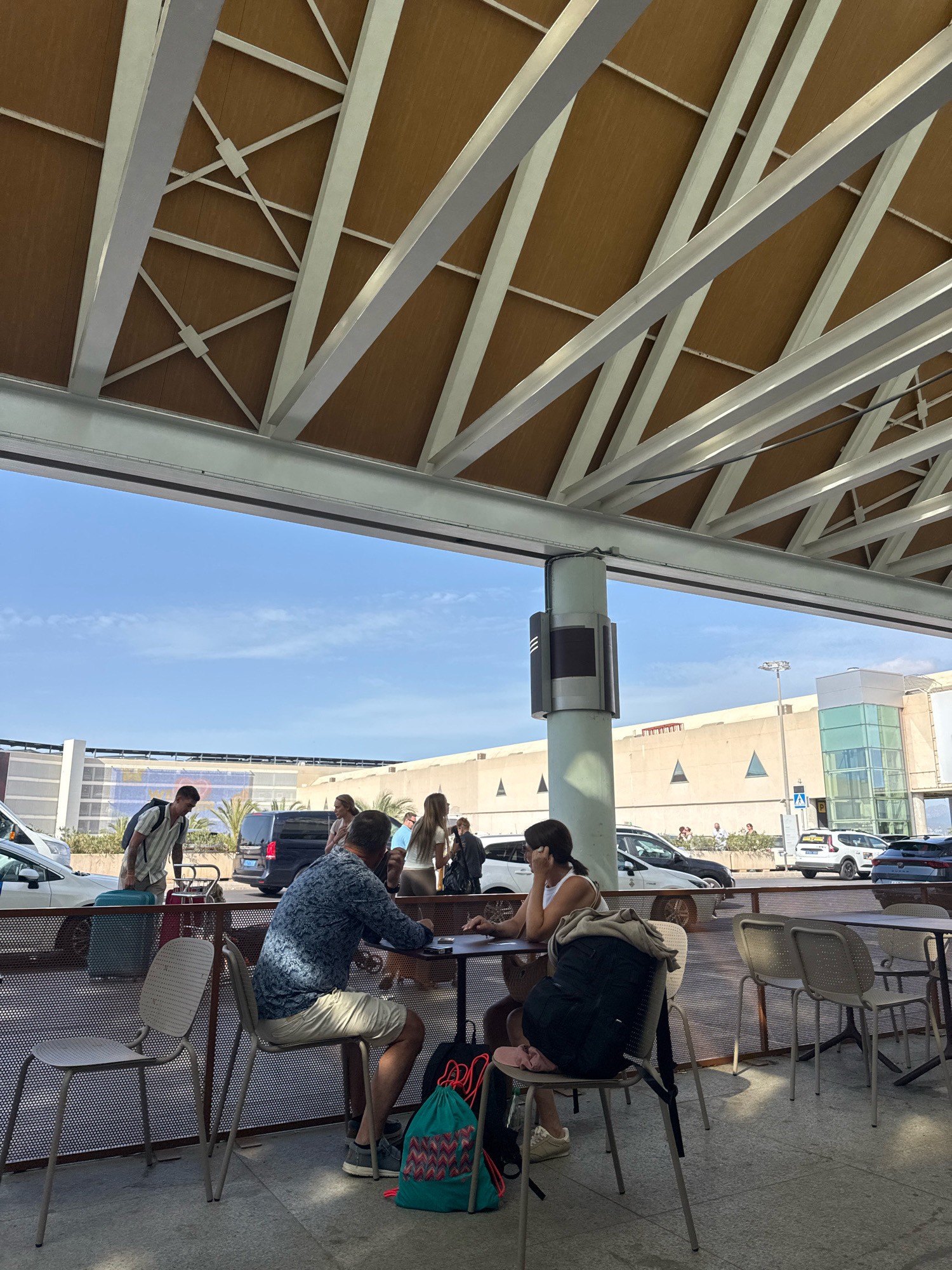 The image captures an outdoor seating area, likely at an airport or transportation hub, in Palma de Mallorca, Spain. Several individuals are present. In the foreground, a man wearing a patterned blue shirt and light-colored shorts sits at a table, appearing to be engaged with a phone or a small object on the table. A woman, wearing a white tank top and shorts, sits opposite him, also looking towards the table. Two chairs are unoccupied at the table. To the left, a man with a backpack and a striped shirt stands, looking towards a dark minivan. Further back, other people are visible near vehicles and buildings, suggesting a departure or arrival area. The environment features a modern architectural style with a large, sloped roof supported by metal beams and columns. Palm trees are visible in the background, hinting at the Mediterranean climate. The sky is clear blue.