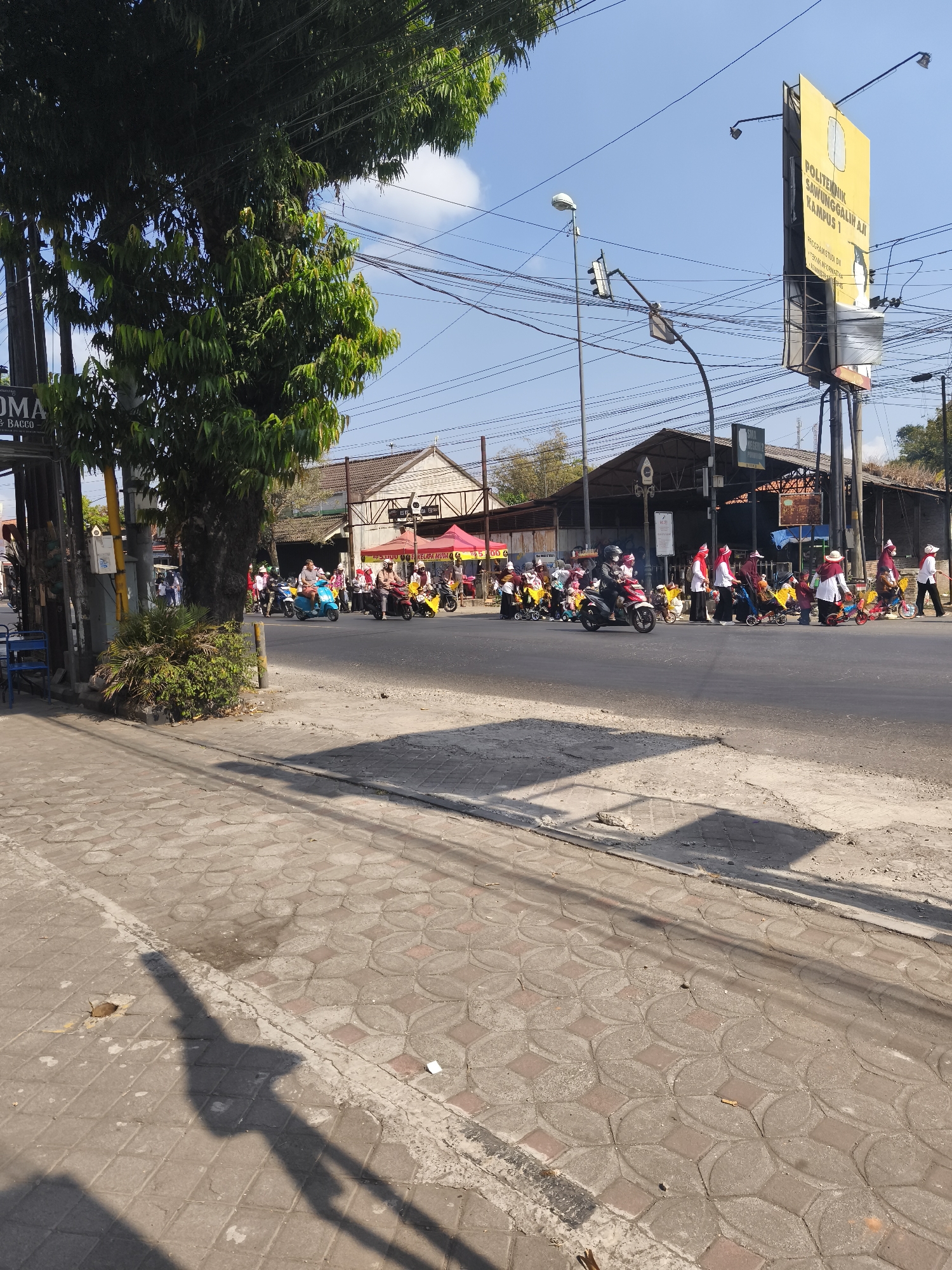 The image depicts a street scene in Kutoarjo, Indonesia, on a sunny day with a clear blue sky. A parade or procession of people on motorbikes and some on foot is moving along the road. The participants are mostly dressed in white and red attire, with many wearing red headscarves or hats. Some individuals are riding scooters, while others are walking alongside, some pushing small carts or what appear to be baby strollers. In the background, buildings with awnings and a tall yellow billboard can be seen. The billboard displays text that includes "POLITEKNIK SAUNGGALIN AJI KAMPUS 1". The road is paved, and a patterned tiled sidewalk is visible in the foreground. Trees, power lines, and street lights are also present in the scene. The overall atmosphere suggests a community event or celebration taking place outdoors.