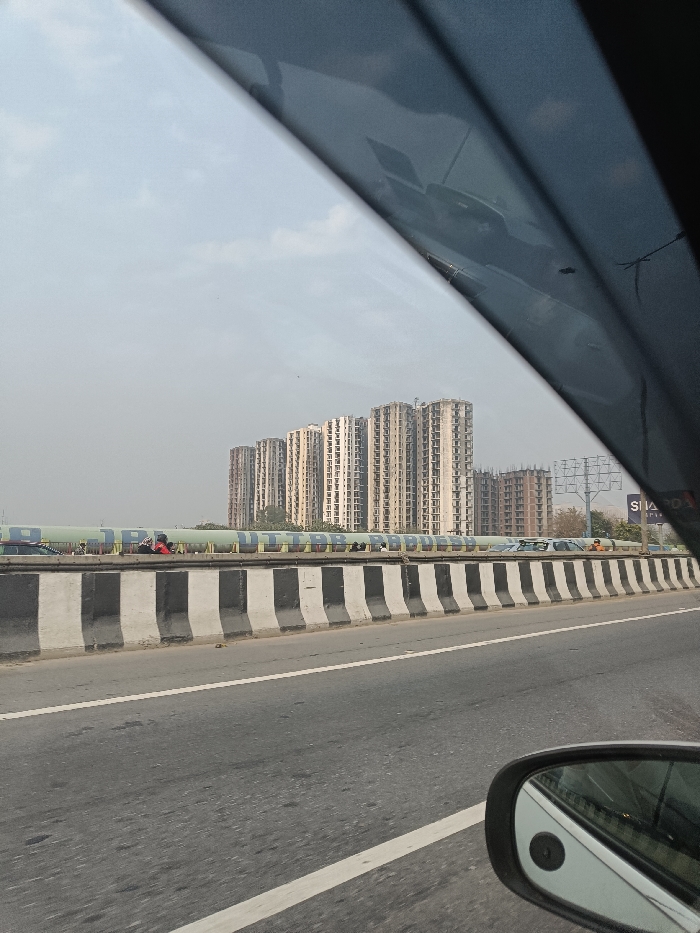 The image captures a view from inside a vehicle driving on a multi-lane highway or expressway under a hazy, overcast sky. In the foreground, the asphalt road features white lane markings. A series of black and white striped concrete barriers separate the active road from a raised median or adjacent structure.
Behind these barriers, a long, light blue or green cylindrical pipeline or covered structure runs horizontally across the mid-ground. Prominently displayed in yellow letters on this structure is the text "UTTAR PRADESH," indicating the geographical location as the Indian state of Uttar Pradesh. A few individuals are visible on or near this structure; one person in a red top and another in a white top can be discerned.
In the background, a cluster of numerous tall, multi-story residential or commercial buildings, appearing to be under construction or recently completed, dominates the skyline. These buildings are uniformly beige or light grey, with many visible windows and structural elements. To the far right, part of a billboard structure is visible, with partially obscured text that seems to include "SI" or "SIA" and "ORP".
The perspective is from the passenger side, with a dark, diagonal obstruction in the top portion of the frame, likely the car's window frame or sun visor. A side-view mirror of the car is visible in the bottom right corner, reflecting a portion of the car's body. The scene depicts an urban or semi-urban environment consistent with Ghaziabad, India, characterized by significant infrastructure and ongoing development during daylight hours.