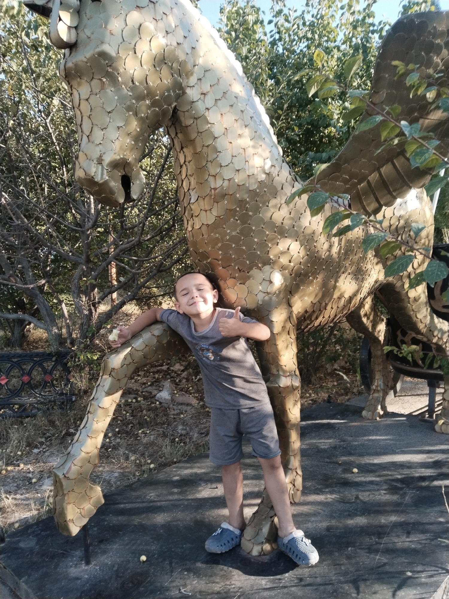 A young boy with short dark hair and a grey t-shirt and shorts is standing next to a large, golden, mythical creature statue. The statue appears to be a dragon or a winged horse, made of numerous overlapping golden scales. The boy is leaning against the statue's leg with his left arm, holding a small piece of food in his hand. His right arm is bent at the elbow, with his thumb up in a "thumbs up" gesture. He is wearing blue slip-on shoes. The statue and the boy are in an outdoor environment, with trees and foliage in the background. The ground is covered in dry grass, fallen leaves, and small yellow fruits. A black metal fence with circular patterns is visible to the left of the boy. The scene suggests a public area or park, possibly in the city of Akata, Uzbekistan, given the presence of the local flora and potentially a local art installation.