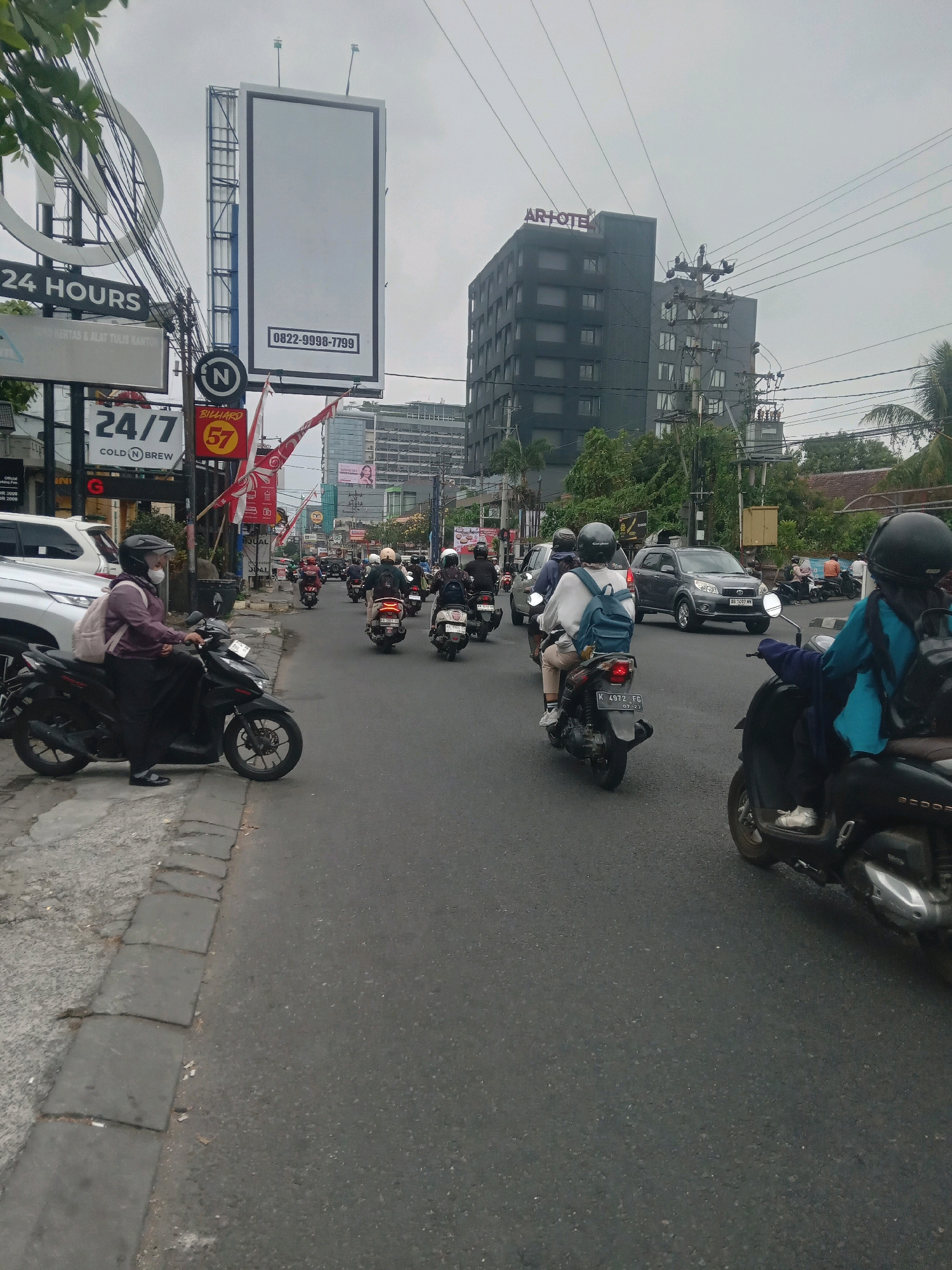 The image shows a street scene in Depok, Indonesia, on a cloudy day. There are many motorcycles and a few cars on the road. Most of the motorcycles are being ridden by people wearing helmets and masks. There are several buildings and billboards visible in the background, including a tall, dark building with "AR HOTEL" written on top and a large billboard with a blank white screen and a phone number. There are also streetlights and power lines overhead. The street is paved with asphalt, and there is a sidewalk on the left side of the road. Some of the motorcycles are parked along the sidewalk, while others are in motion on the road. The overall scene depicts a busy urban environment with active traffic.