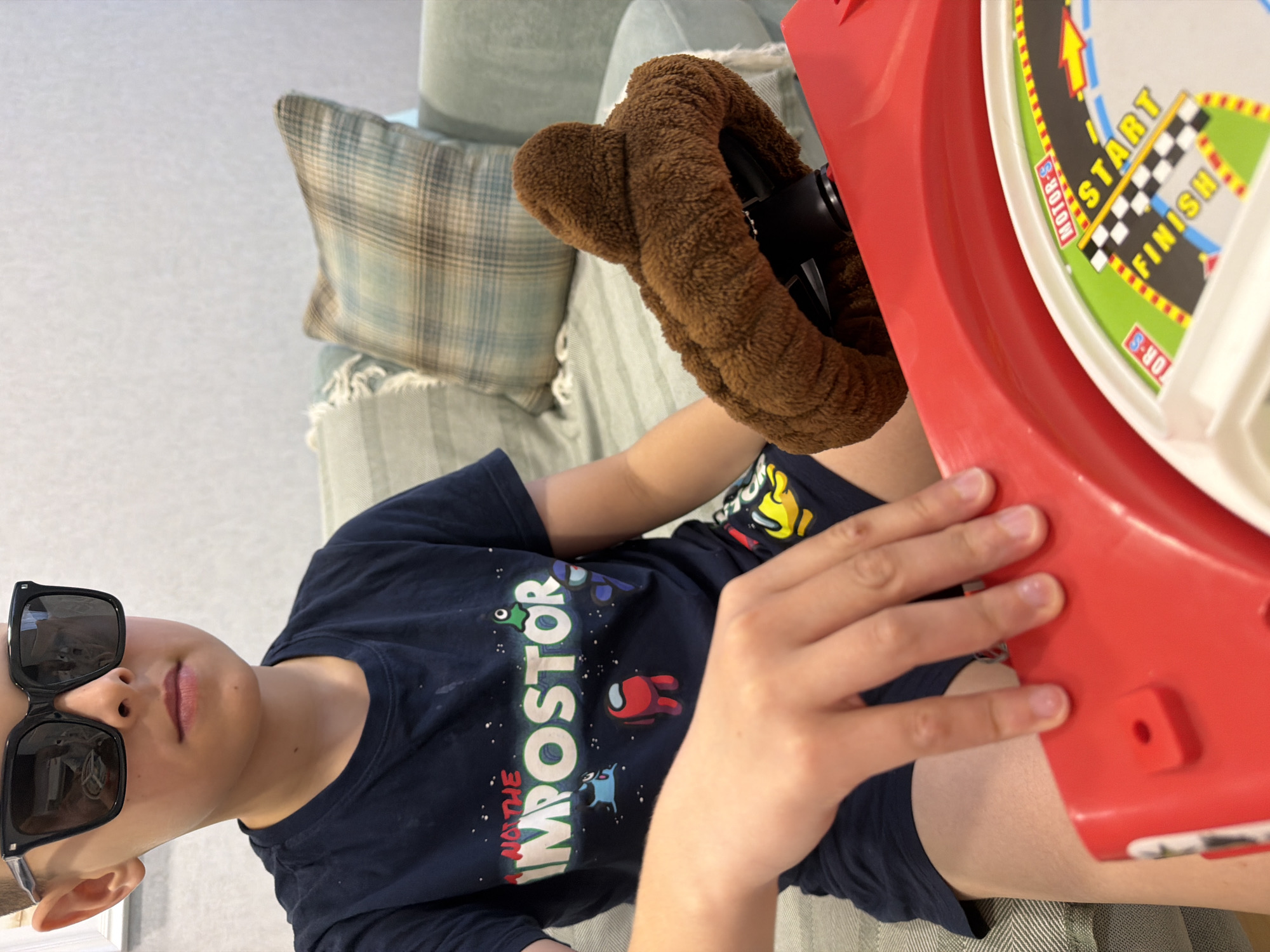 A child wearing sunglasses and a blue "Not the Impostor" t-shirt is seated on a light green couch with a plaid throw pillow. They are holding a red toy race car with a brown steering wheel cover. The toy race car has "Start" and "Finish" written on it. The location is unknown, and there is no indication that this image was taken in Votkinsk, Russia.