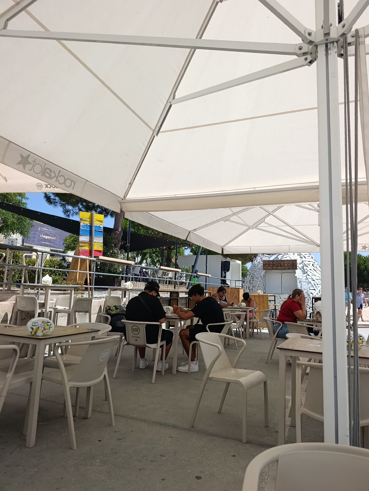 An outdoor seating area of a restaurant is depicted in Setúbal, Portugal. The scene is set under a large white canopy, supported by metal structures. Several tables with white chairs are arranged on a concrete floor. A few people are seated at the tables, some eating and others conversing. In the background, there are trees, a building with a prominent colorful wooden sculpture, and a beach with people walking. The overall atmosphere suggests a casual dining experience in a sunny, urban environment.