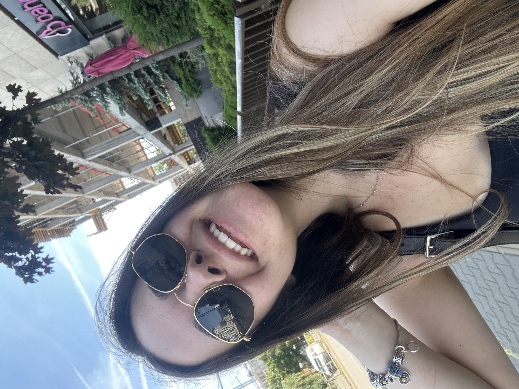 A selfie of a young woman with long, light brown hair and sunglasses is taken outdoors in Sofia, Bulgaria. The woman is smiling broadly and looking at the camera. She is wearing a black tank top and a silver bracelet with charms. In the background, a building under construction is visible, along with trees and a pink umbrella. The reflection in her sunglasses shows a street with buildings.