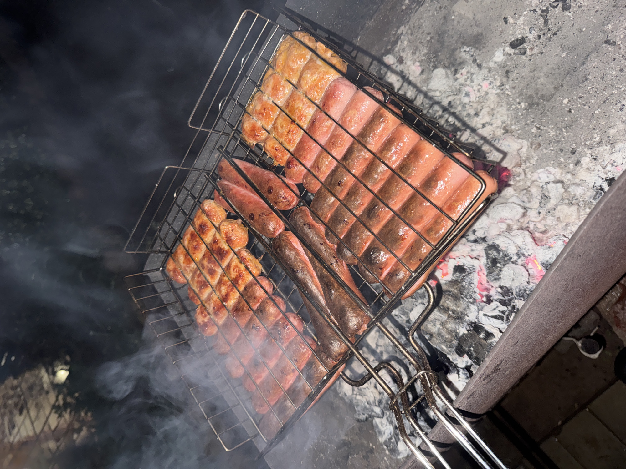 The image shows a grill basket containing sausages and patties being cooked over hot coals. The grill basket rests on the edge of a concrete fire pit. Visible in the lower left corner is smoke, presumably from the burning coals. The context of the city of Mayak, Russia is not evident in the image.
