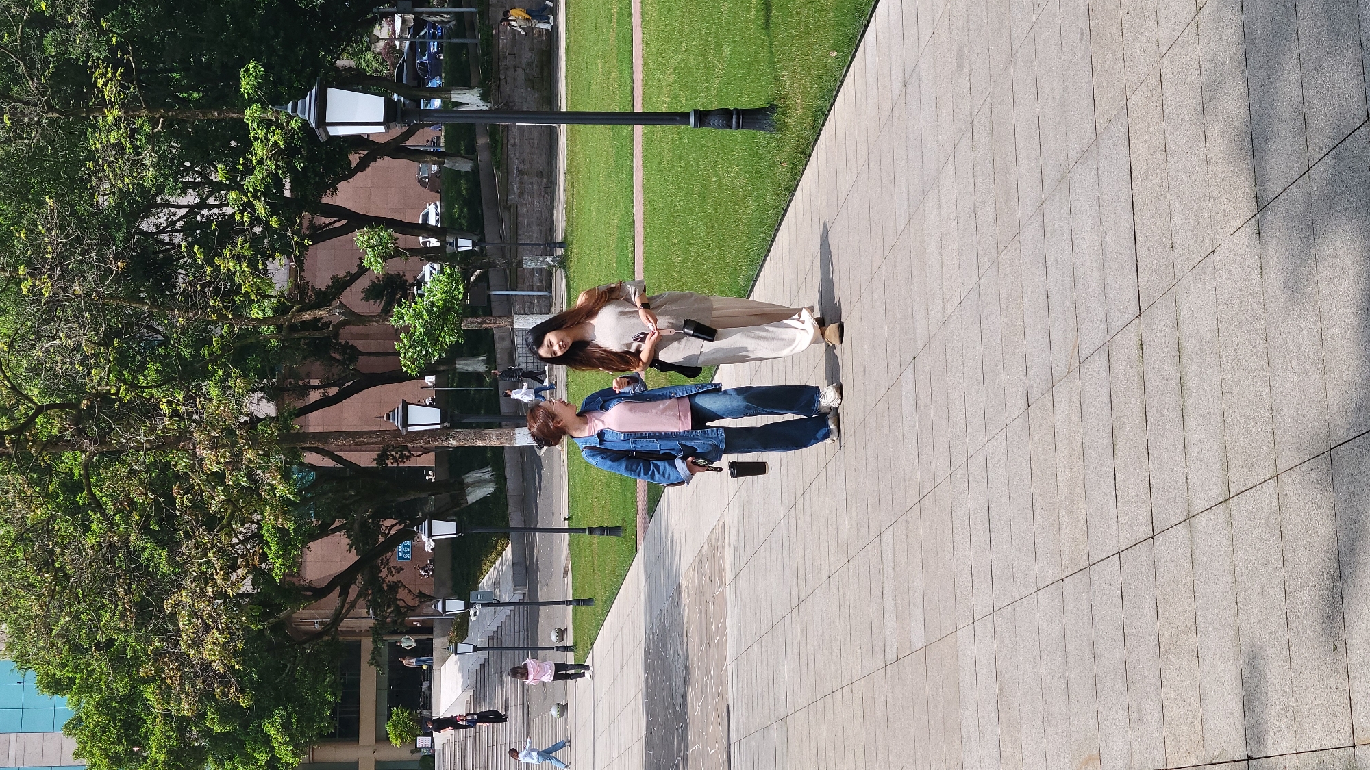 Two women are standing together on a tiled walkway with green lawn to their left. The woman on the left wears a denim jacket over a pink shirt and jeans. The woman on the right wears light-colored pants and a long-sleeved top. Both women appear to be holding small, dark-colored objects in their hands.
Behind them, a line of streetlights stands alongside a row of trees. In the background, other people can be seen walking. The buildings and scenery suggest an urban setting.