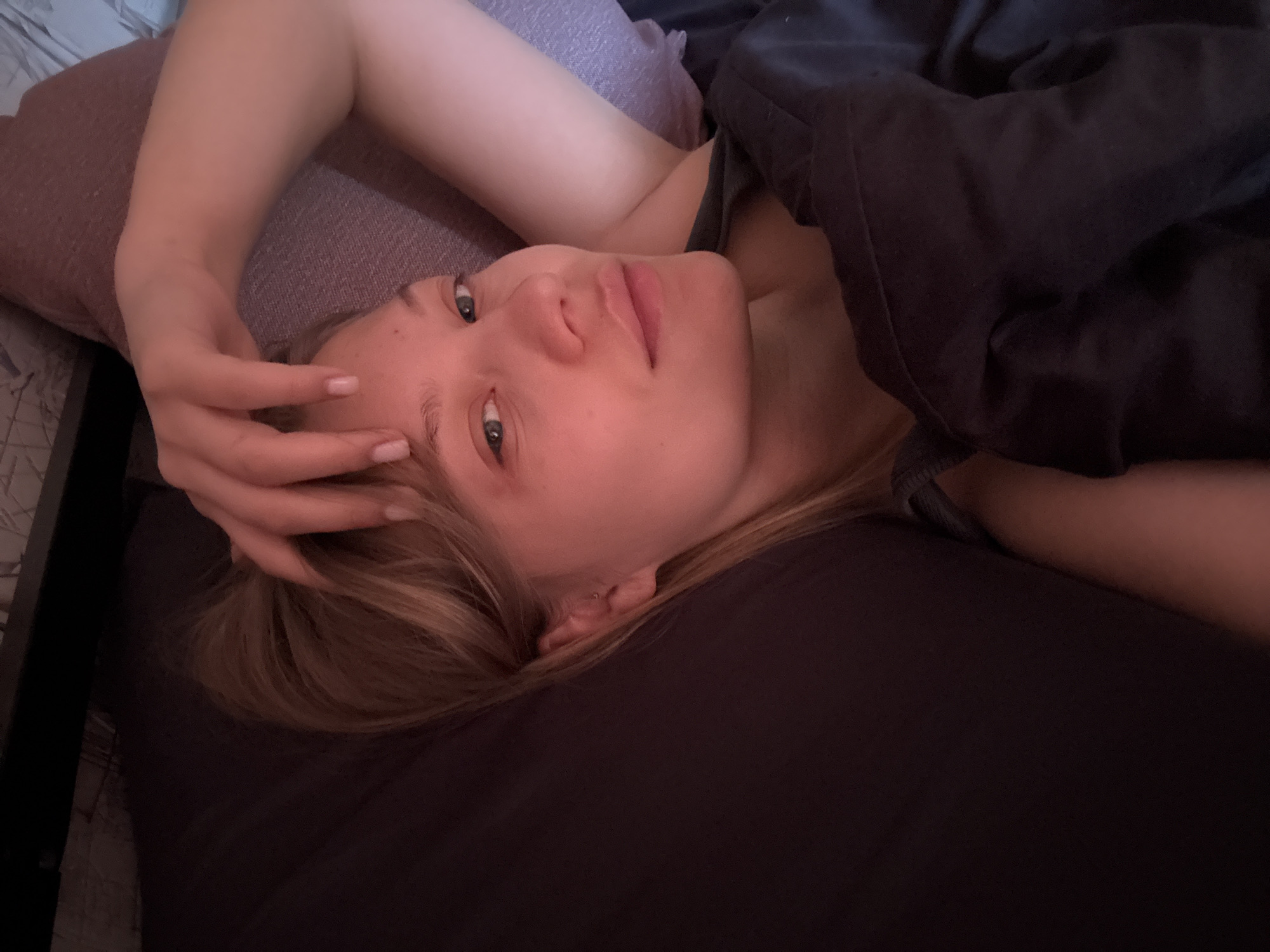 A woman is lying in bed, resting her head on a pillow and her hand on her forehead. She has blonde hair, fair skin, and light eyes. She is partially covered by a dark-colored blanket. The background shows a partially visible headboard. There is no indication of the location being Chaikovsky, Russia.