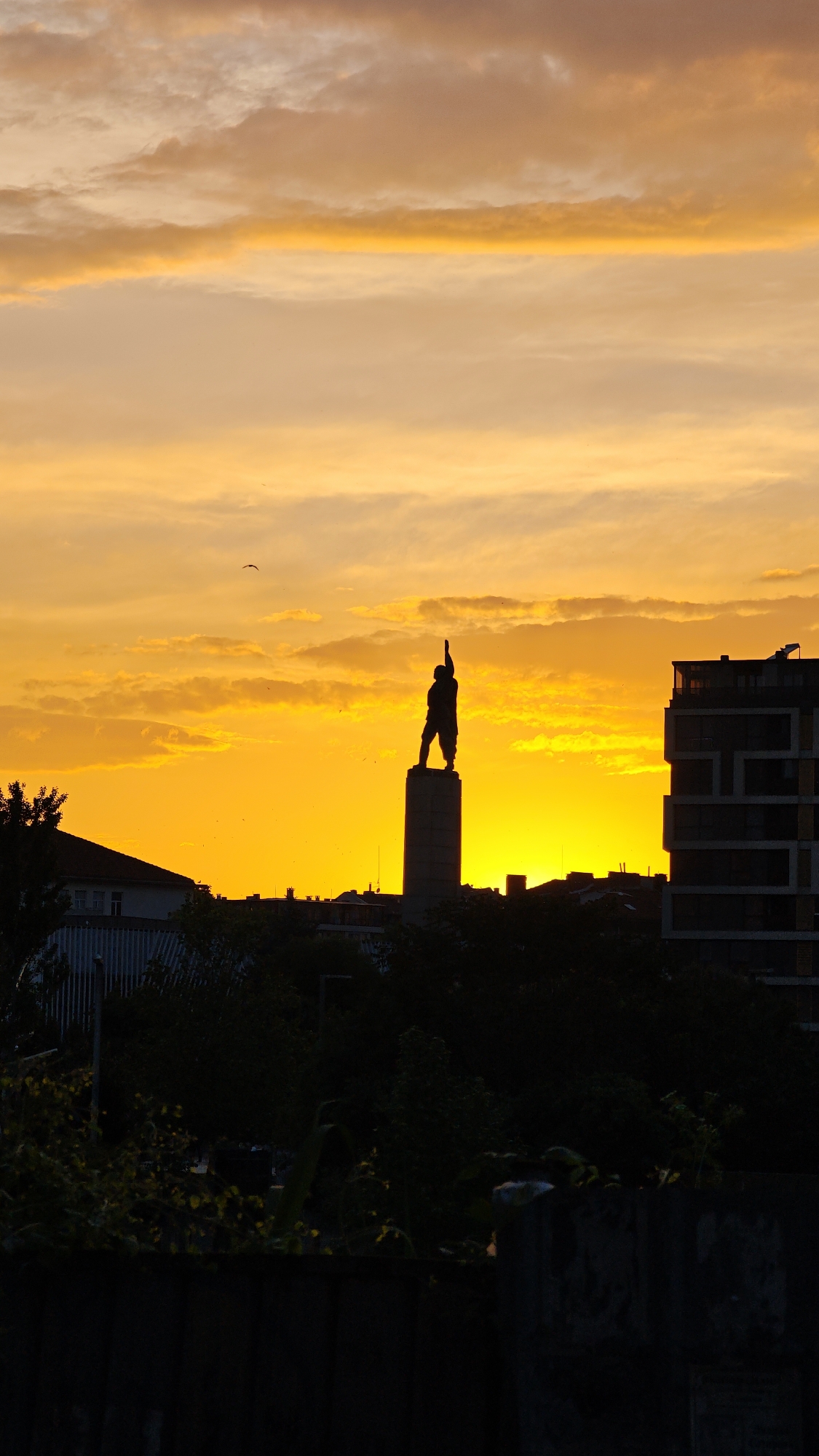 The image shows the Alyosha monument in Burgas, Bulgaria silhouetted against a vibrant golden and orange sunset. The statue of the Soviet soldier stands atop a cylindrical pedestal. The foreground is dark, with a fence and greenery obscuring the view. In the background, buildings and trees are visible, also in silhouette. There is a bird in flight in the sky.