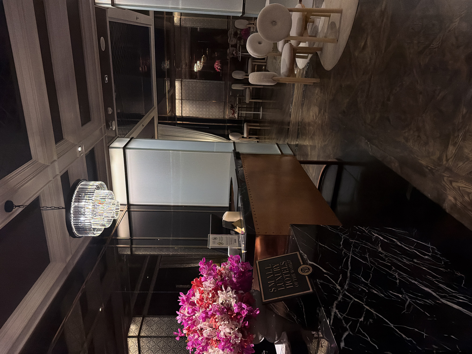 The image shows the reception area of the The Athenee Hotel, a Luxury Collection Hotel, Bangkok. The reception desk is made of black marble with a copper finish and is adorned with a large bouquet of pink and white orchids. A sign on the desk reads "SMALL LUXURY HOTELS OF THE WORLD". In the background, there are several chairs arranged around small tables, with a chandelier hanging from the ceiling. The hotel's ornate ceiling design and the polished floor contribute to the luxurious ambiance.
