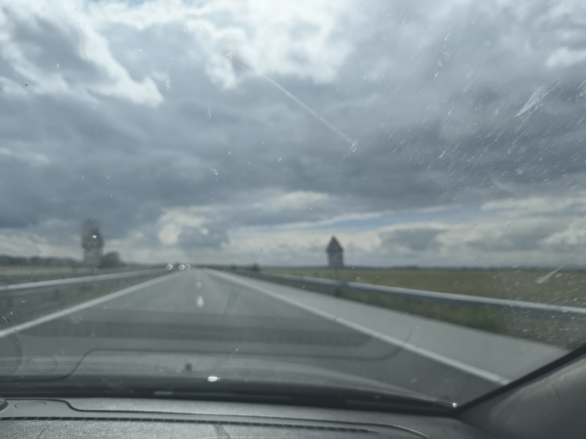 The image shows a view from inside a car, looking through the windshield at a highway on a cloudy day. The highway stretches into the distance, with white lines marking the lanes. On the right side of the highway, there is a field and a guardrail. In the distance, there is a building with a distinctive roof shape, possibly a historical landmark. The sky is filled with gray clouds, suggesting an overcast or impending weather event. The dashboard of the car is visible in the foreground. There are smudges and specks on the windshield, which obscure the view slightly. The location is identified as the city of Zheleznik, Bulgaria.