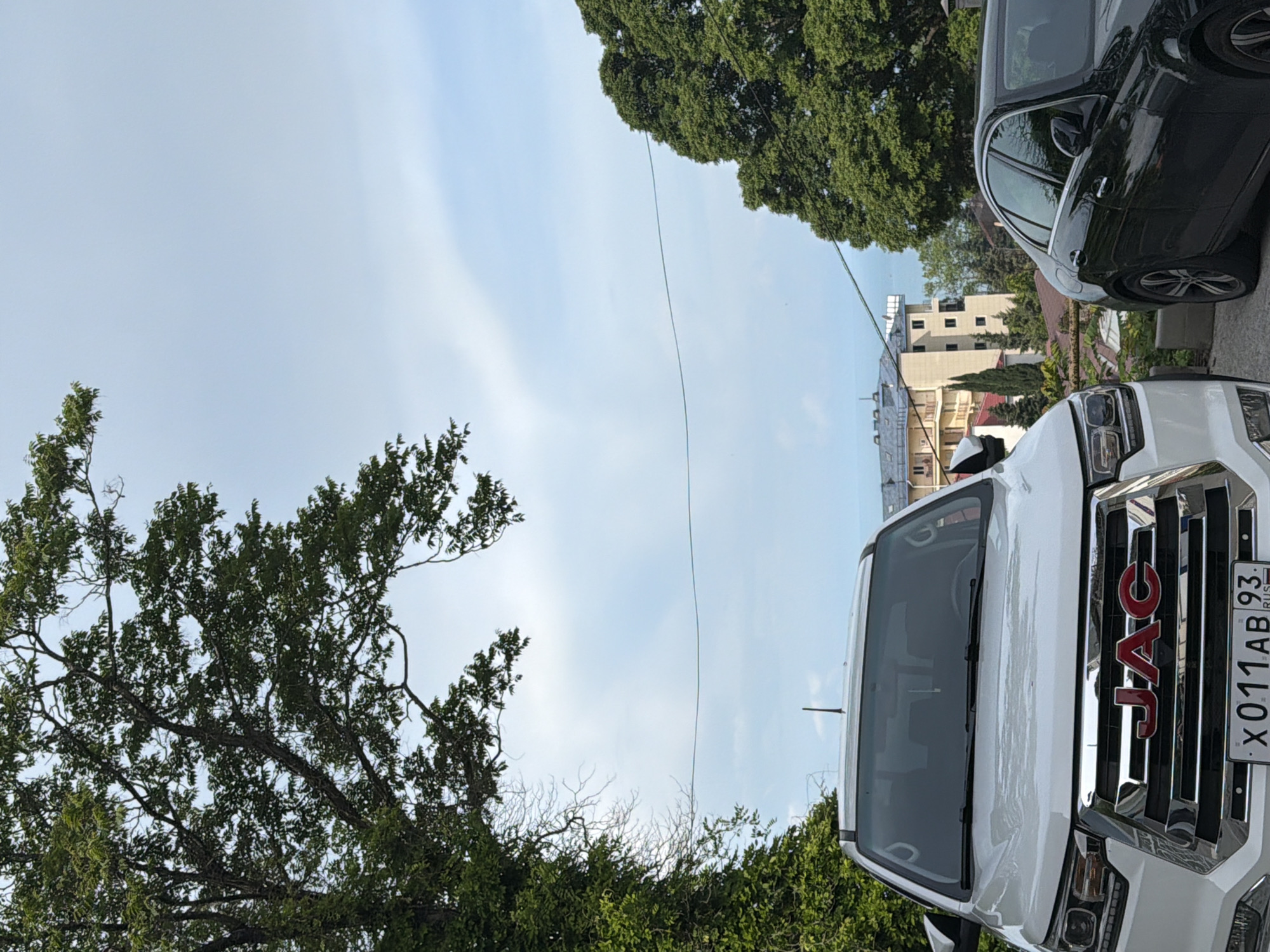 In Sochi, Russia, two cars are parked on a slight incline under a bright, overcast sky. To the left, a white JAC SUV dominates the foreground, its chrome grille and red JAC logo clearly visible. The license plate reads "X011AB 93 RUS". To the right, a black sedan is partially visible, with its rear passenger door, window, and part of the wheel showing. In the background, a multi-story building with a distinctive cream-colored facade and blue-tiled roof sits amongst lush green trees and foliage. A sliver of the sea can be seen between the building and the trees on the right. The scene is framed by the branches of a large, leafy tree on the left and another dense green tree on the right. Power lines are strung across the sky.