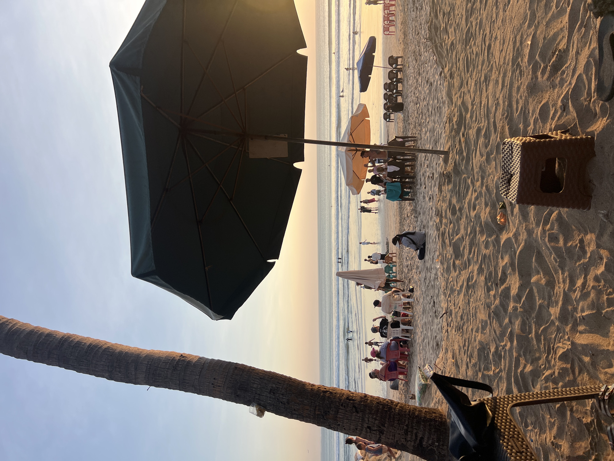 The image depicts a beach scene in Kuta, Indonesia, during sunset. A tall palm tree is visible on the left, with its trunk textured and a piece of white plastic attached near the top. A large, dark green umbrella dominates the foreground, casting a shadow over the sandy beach. In the middle ground, people are seated at tables under a lighter-colored umbrella, engaged in conversation. A few individuals are standing and walking around the area. Further towards the sea, several people are seen near the water's edge, with some individuals swimming or surfing in the ocean. The sea is calm with gentle waves, and the sky is a gradient of pale blue and warm orange hues, indicating the setting sun. Beach chairs and tables are scattered across the sand, some occupied and others empty. The sand itself is textured with ripples.