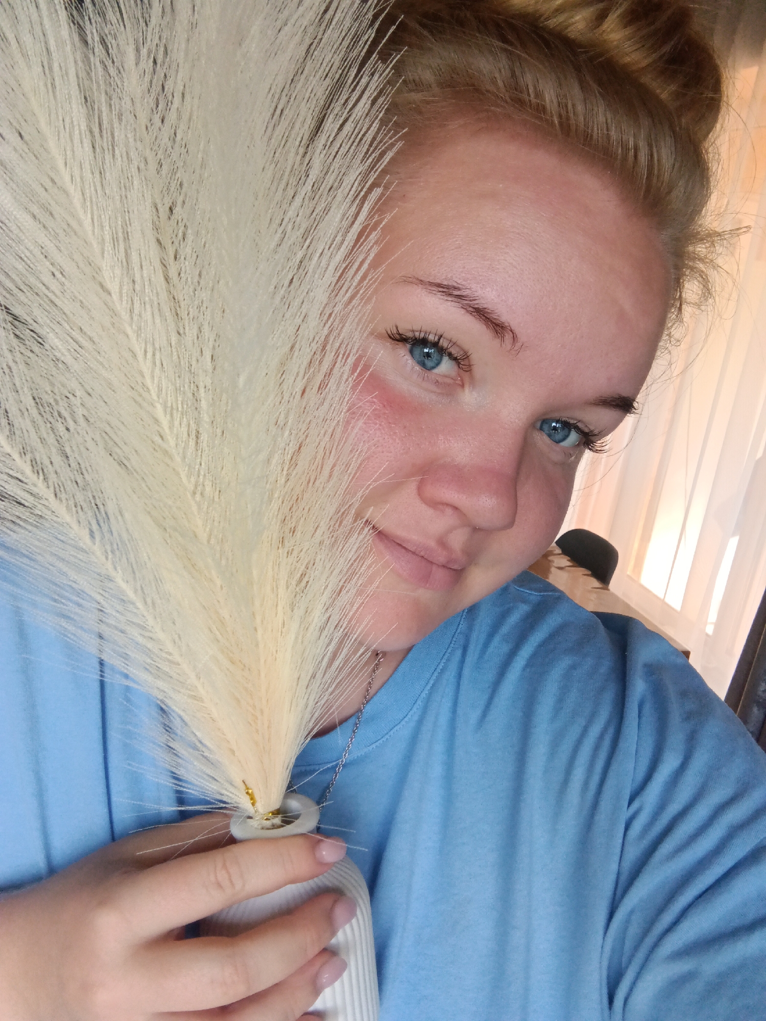 A close-up shot shows a young woman with blonde hair pulled up in a bun. She has bright blue eyes and fair skin. She is wearing a light blue t-shirt and holding a fluffy white plant in a small white vase. The plant obscures part of her face. The background is blurry and appears to be an indoor setting with a window and curtains. There is no indication of the location being the city of Фоки, Russia.