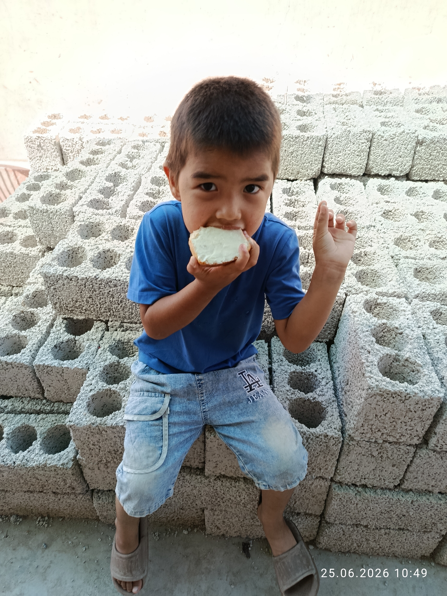 A young boy with dark hair and brown eyes is sitting on a stack of concrete blocks, eating a piece of bread. He is wearing a blue t-shirt and denim shorts. The blocks are gray and appear to be rough and porous. The background is a wall that is lighter in color than the blocks, with some faint markings or discoloration. The overall scene is outdoors, with natural lighting. There is a timestamp in the lower right corner of the image that reads "25.06.2026 10:49".