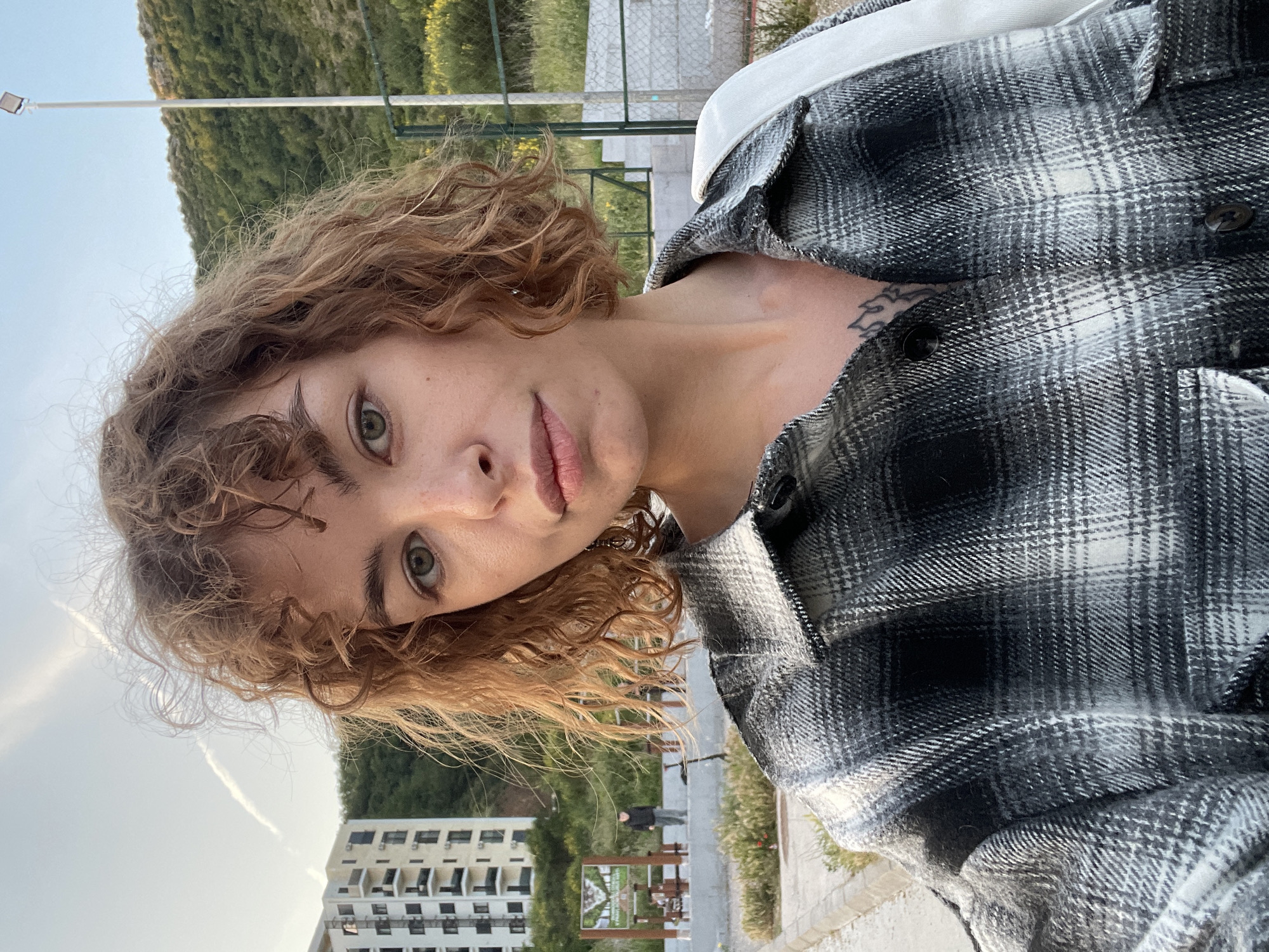 A young adult woman with curly, light brown hair and green eyes is pictured in a selfie. She is wearing a black and white plaid shirt. A tattoo is visible on her upper chest. The background shows an outdoor environment with a building, trees, and what appears to be a walkway. A sign is also visible, potentially indicating a park or scenic area. The location is Boreti, Montenegro, as indicated by the sign on the walkway.