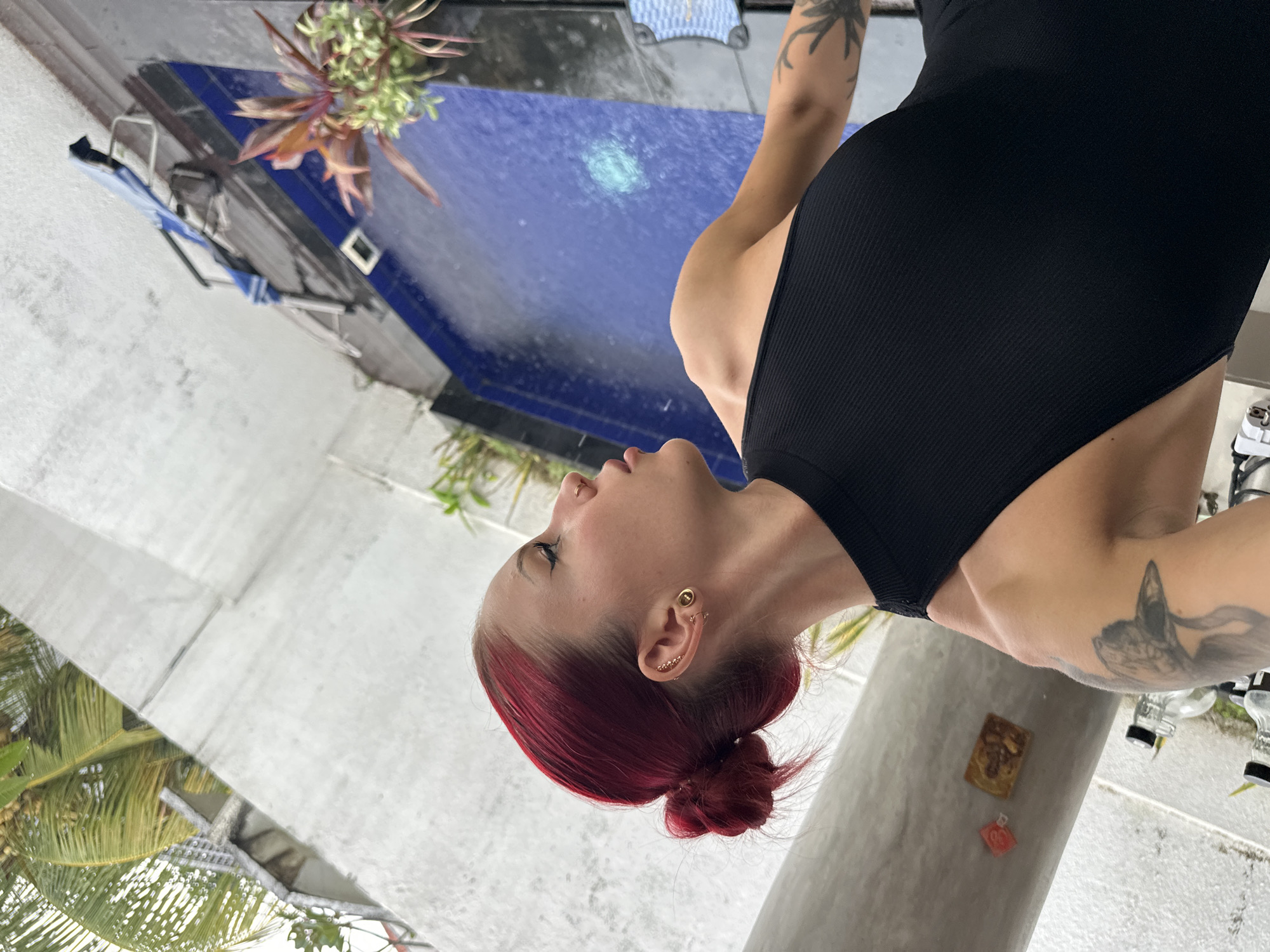 This image captures a woman in an outdoor tropical setting, likely a poolside or patio area in North Kuta, Indonesia. The woman, positioned in the foreground, has vibrant red hair styled into a low bun and fair skin. She is wearing a form-fitting black ribbed sleeveless bodysuit or swimsuit. Her head is tilted slightly upwards and to her left, with her eyes appearing closed or downcast, conveying a serene or contemplative mood. She has multiple gold-toned ear piercings on her visible right ear and a nose piercing. Tattoos are visible on her right arm, including what appears to be a butterfly design near her shoulder.
In the background, a clear blue swimming pool with water ripples occupies a significant portion of the scene. Surrounding the pool are light-colored concrete or paved surfaces. Green tropical foliage, including prominent palm fronds, is visible, indicating a lush environment. Near the pool's edge, a light blue foldable chair and a blue and white patterned mat or cushion can be seen. To the right of the woman, a grey concrete pillar features a small, ornate rectangular plaque-like item and a smaller red square object. Transparent bottles are partially visible in the far right background. The scene is brightly lit, suggesting it is daytime with clear weather. No discernible text is visible.