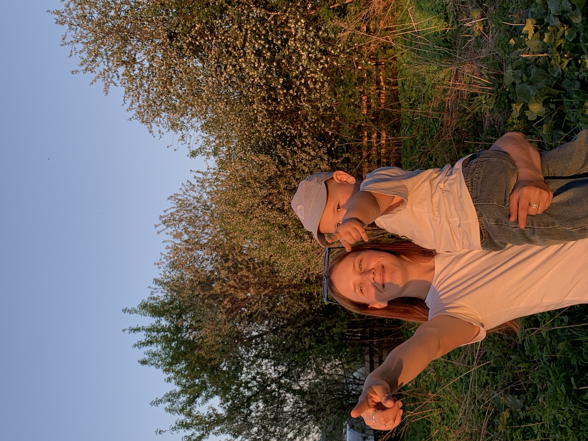 A woman and a young child stand in a grassy area with trees and bushes; the sky is visible in the upper left corner. The woman has reddish-brown hair and wears a white t-shirt. The child, who is on the woman's back, wears a blue cap and a light-colored shirt. Both are pointing their fingers. The location appears to be outdoors, in an open green space. There is no indication of whether the location is in the city of Фоки, Russia.