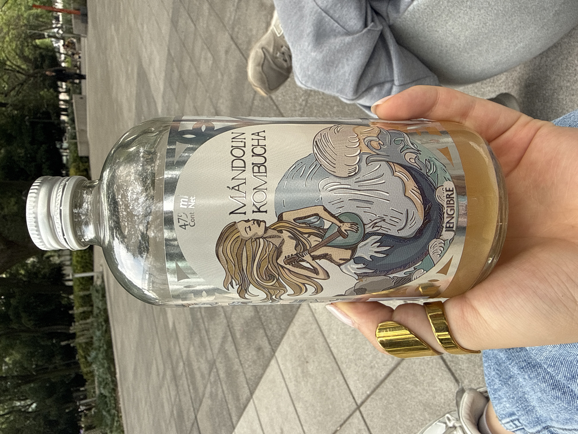 A glass bottle of Mandolin Kombucha, labeled "Mándolin Kombucha Jengibre," is held by a person wearing a gold ring. The bottle is partially filled with a light brown liquid. The background shows a paved area with trees, suggesting an outdoor location. There are people visible in the background, implying a public space. The style of the paving suggests the location is in Mexico City, Mexico.
