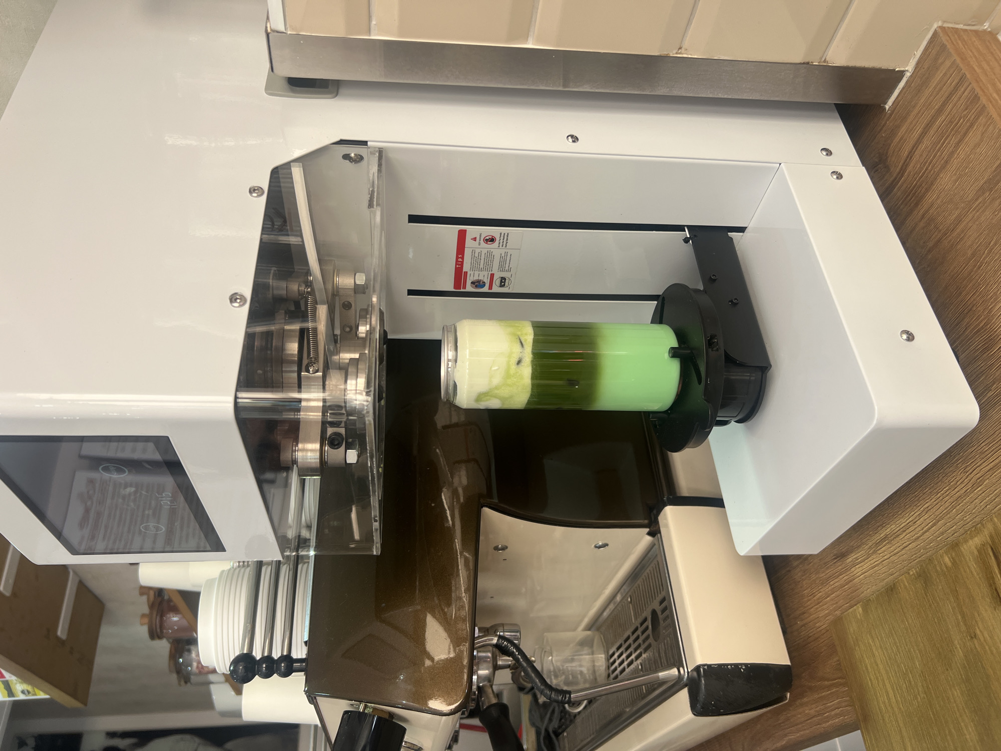 A beverage cup containing a green and white liquid is mounted within a specialized machine. The machine features a white exterior, a transparent protective shield, and various mechanical components including visible bolts and metallic gears. Parts of an espresso machine are visible below. The background consists of tiled walls and wooden cabinetry. The specific location in London is indiscernible without additional contextual clues.