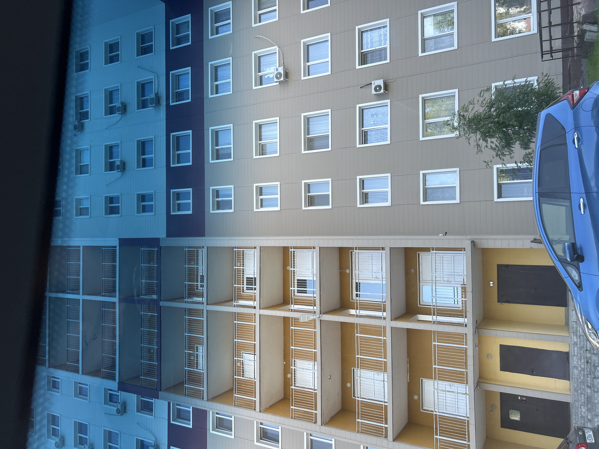 The image shows a modern apartment building in Orenburg, Russia. The building is tall, with multiple stories featuring balconies and windows. The facade is a mix of grey and yellow siding, with white railings on the balconies and white window frames. Several air conditioning units are visible on the exterior walls.
In the foreground, a blue car is parked on the street in front of the building. The car is facing towards the right side of the image. The building's entrance is visible on the ground floor, with several dark doors. The environment appears to be a residential area, with some greenery and trees visible around the building. The sky is not clearly visible due to the angle of the shot and possible reflections.
