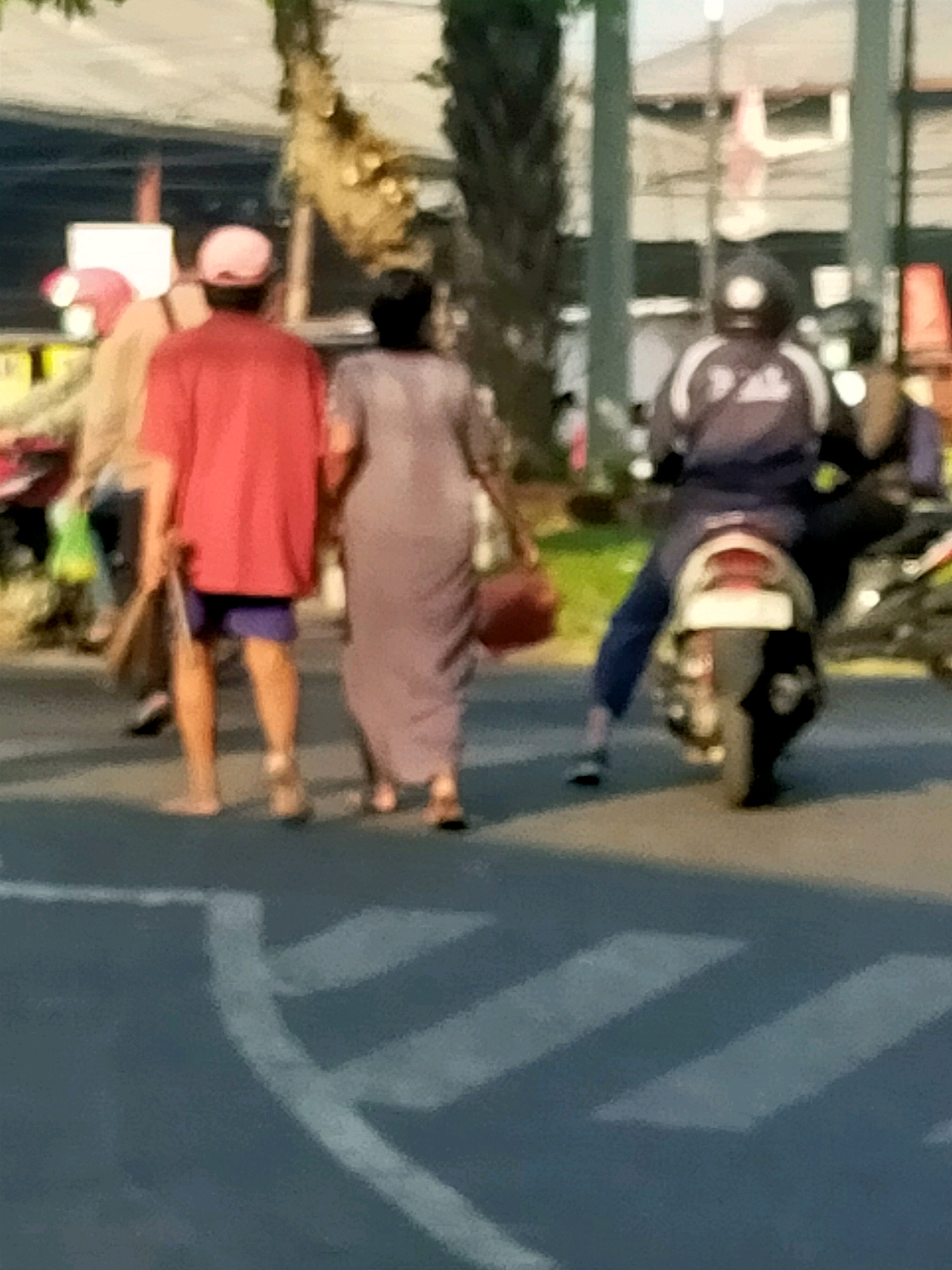 The image depicts a street scene, likely in Surabaya, Indonesia, with a focus on pedestrians and a person on a motorcycle. In the foreground, a zebra crossing is visible on the asphalt road. Three individuals are walking away from the viewer. The leftmost person is partially obscured, wearing a light-colored garment. The person next to them, in the center, is wearing a bright red t-shirt and dark blue shorts, carrying a brown bag. The third person, to the right, is wearing a long, light-colored dress and carrying a reddish-brown bag. Further in the background, a person is riding a motorcycle, wearing a dark helmet and a dark uniform with white accents. The environment suggests an urban setting with buildings and trees in the background, illuminated by natural light, indicating it is daytime.