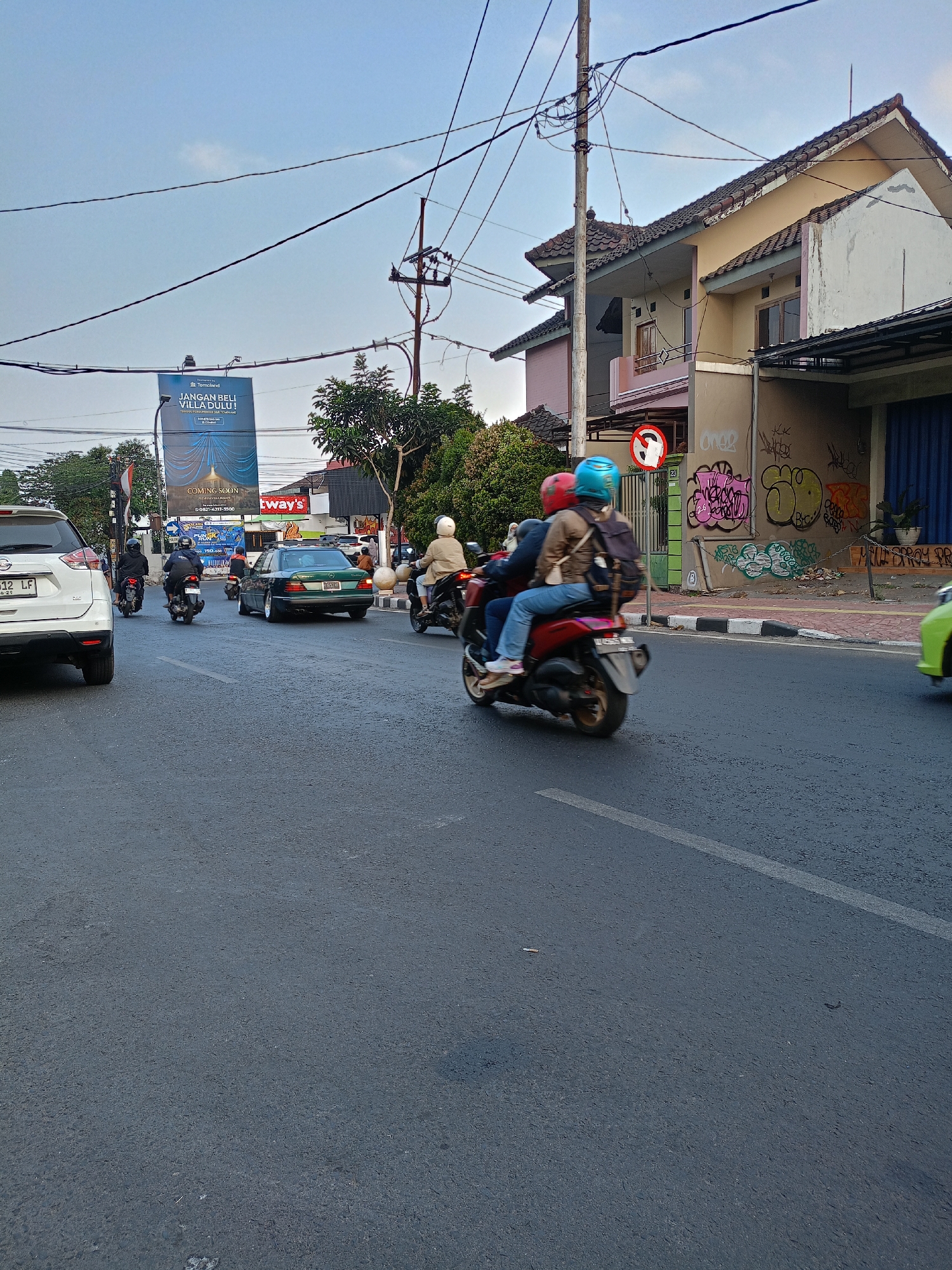 The image depicts a street scene in Batu, Indonesia, likely during daylight hours given the clear sky and moderate lighting. A busy road is the focal point, with several vehicles present. On the left, a white SUV is parked, its license plate clearly visible. Further down the road, a green sedan is in motion, and multiple motorcycles are also navigating the street. Two individuals are riding a red scooter in the foreground, with the passenger wearing a red helmet and the driver a blue helmet. Their clothing is casual, suggesting everyday activity.
The urban environment is characterized by buildings lining the street, some appearing to be residential homes with tiled roofs and balconies. Graffiti is visible on the wall of one building. A large billboard advertising a villa complex is prominently displayed on the left side of the road, with text in Indonesian, including "JANGAN BELI VILLA DULU!" which translates to "Don't buy a villa yet!". This suggests a commercial or residential development being promoted. Overhead power lines crisscross the sky, a common feature of urban infrastructure. The road surface appears to be asphalt, with visible lane markings. The overall impression is that of a typical day in a moderately busy Indonesian city.