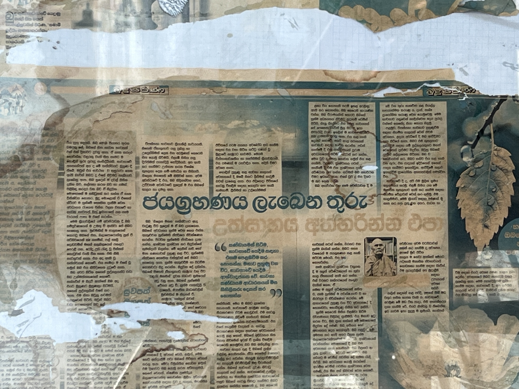 The image depicts a close-up, overhead view of a collage or mixed-media artwork that incorporates elements of vintage newspapers, distressed paper, and possibly photographic transfers. The dominant visual is a collection of text in what appears to be Sinhala script, printed on aged, yellowed newspaper pages. There are areas of the paper that are stained with brown, resembling water damage or coffee stains, and some sections are overlaid with a semi-transparent white material, possibly paint or a wash.
Towards the right side of the image, there is a large, stylized heading or title in bold, dark green Sinhala script. Below this, running vertically, is another title in a similar script. To the left of these titles, two quotation marks are visible, suggesting the presence of quoted text within the newspaper.
In the lower middle section of the image, a small, sepia-toned photograph is embedded within the collage. The photograph shows an elderly man with a beard, wearing glasses and a dark shirt, seated at a desk with papers in front of him. He appears to be looking slightly off-camera.
The background of the artwork consists of layers of aged paper, with some areas exhibiting a subtle green hue, potentially from a printed image or a colored overlay. There are also hints of photographic imagery or artistic renderings of natural elements, such as a leaf and possibly a landscape at the bottom. The overall aesthetic is one of decay, history, and artistic layering.
The provided information states the location is the city of Matara, Sri Lanka. While the image itself does not contain explicit geographical markers of Matara, the presence of Sinhala script strongly suggests a Sri Lankan context. Without further information or visual cues directly referencing Matara, its specific inclusion in the description is based solely on the provided prompt.