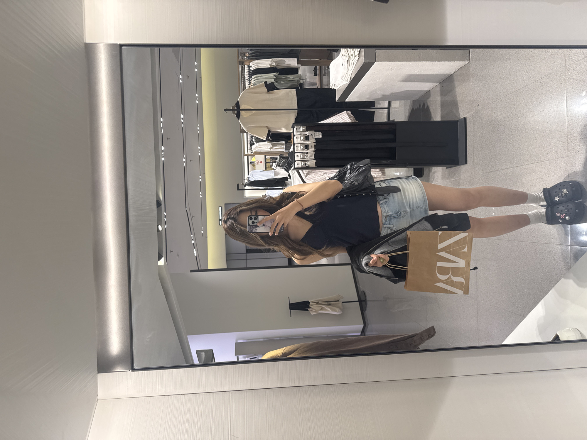 A female is reflected in a mirror inside a retail store. She is holding a mobile phone up in her right hand, seemingly taking a photo of herself. She is wearing a dark top, a denim skirt, white socks, and Crocs. She carries a black purse and a brown shopping bag labeled "ZARA". The store setting shows racks of clothing, display tables, and a bright, tiled floor. The mirror's frame is visible. It is not possible to determine if this image was taken in Yuzhong District, China.