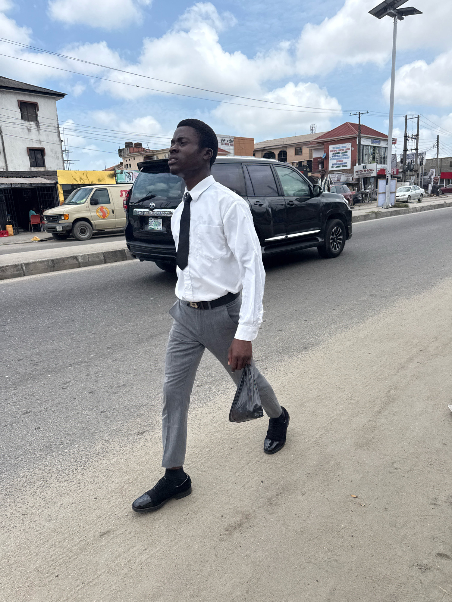 A young Black man, dressed in a white button-down shirt, black tie, and grey dress pants, walks across a paved road in Ado, Nigeria. He carries a black plastic bag in his right hand and wears black dress shoes. The man is looking up and to his left. Behind him, a black SUV is parked on the side of the road, and a beige van is visible further down the street. In the background, buildings and utility poles line the street under a partly cloudy sky. The overall scene depicts a street in a busy urban environment.