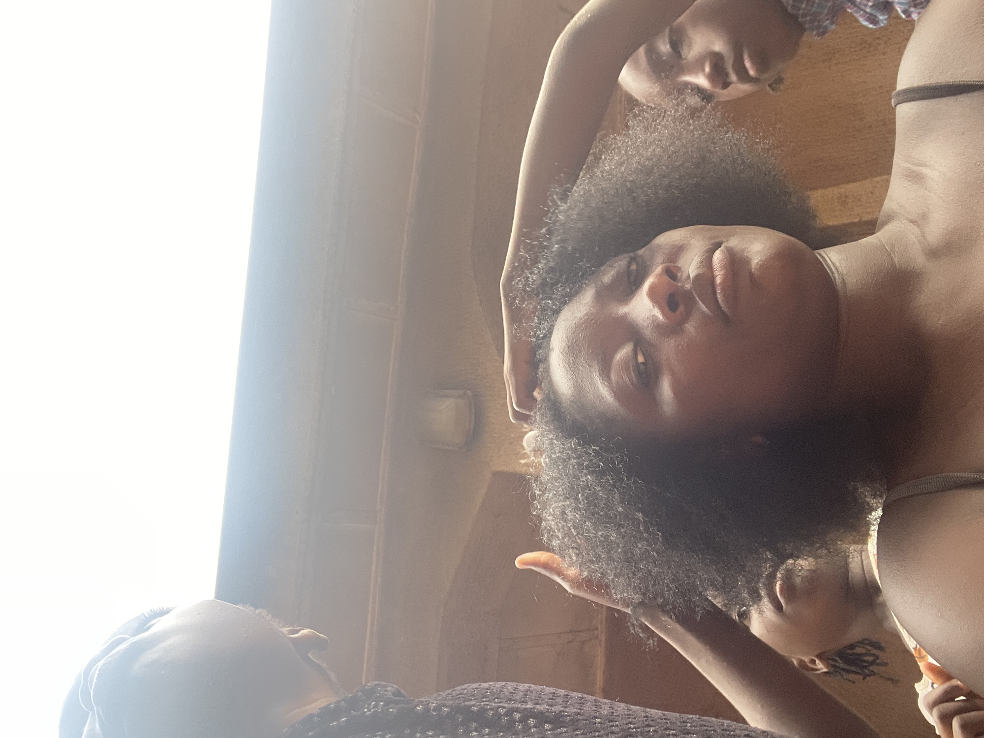 The image, captured from a low angle looking upwards, features three individuals in a close-up composition. The dominant subject is a person with dark, curly afro-textured hair, positioned in the center-right, looking directly down towards the camera with a neutral expression. They wear a light-colored top, possibly a tank top, with a dark strap visible on their right shoulder. Their skin tone is dark.
Behind this main subject, in the upper right portion of the frame, another individual's face is partially visible, also looking downwards. This person appears to be wearing a plaid or checked garment. A third individual is partially seen in the lower left foreground, with their back and the side of their head facing the camera as they look towards the left. This person also has dark, curly hair and is wearing a dark, possibly sparkly, textured top.
The background consists of a warm-toned, textured wall, possibly brick, which extends across the right side and upper portions of the image. A light fixture is visible on this wall in the mid-ground. The left side of the image is dominated by an extremely bright, almost white light source, suggesting a window or open doorway allowing ample daylight into the space. The scene appears to be an indoor or shaded outdoor setting. The bright light suggests it is daytime. No discernible text is visible in the image. The overall atmosphere is casual and relaxed. Given the context, the setting is Akwanga, Nigeria.