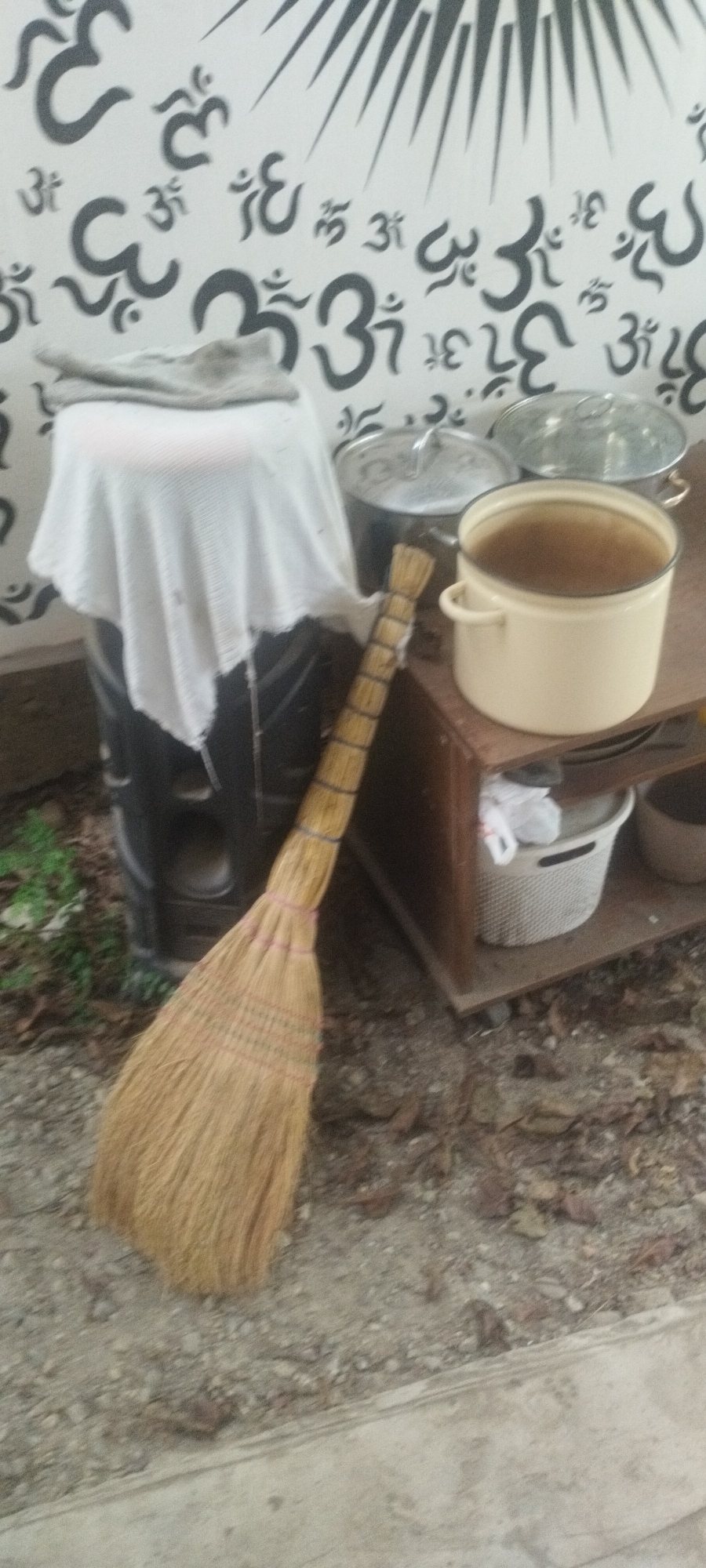 An outdoor, medium-shot, eye-level photograph shows a rustic scene with an emphasis on traditional elements. In the background, a wall is decorated with black Om symbols and a sunburst design. In the mid-ground, a large black cylindrical object, possibly a barrel or a stove, is partially covered by a white cloth. To the right of this object, on a wooden shelf, are two metal pots with lids, and a larger, cream-colored pot filled with a brownish liquid. Leaning against the black object and extending into the foreground is a broom made of straw, tied with pink and yellow string. The ground is covered with dirt, small rocks, and scattered leaves, suggesting a natural or semi-outdoor setting. The overall impression is one of a quaint, possibly rural or bohemian, environment.
