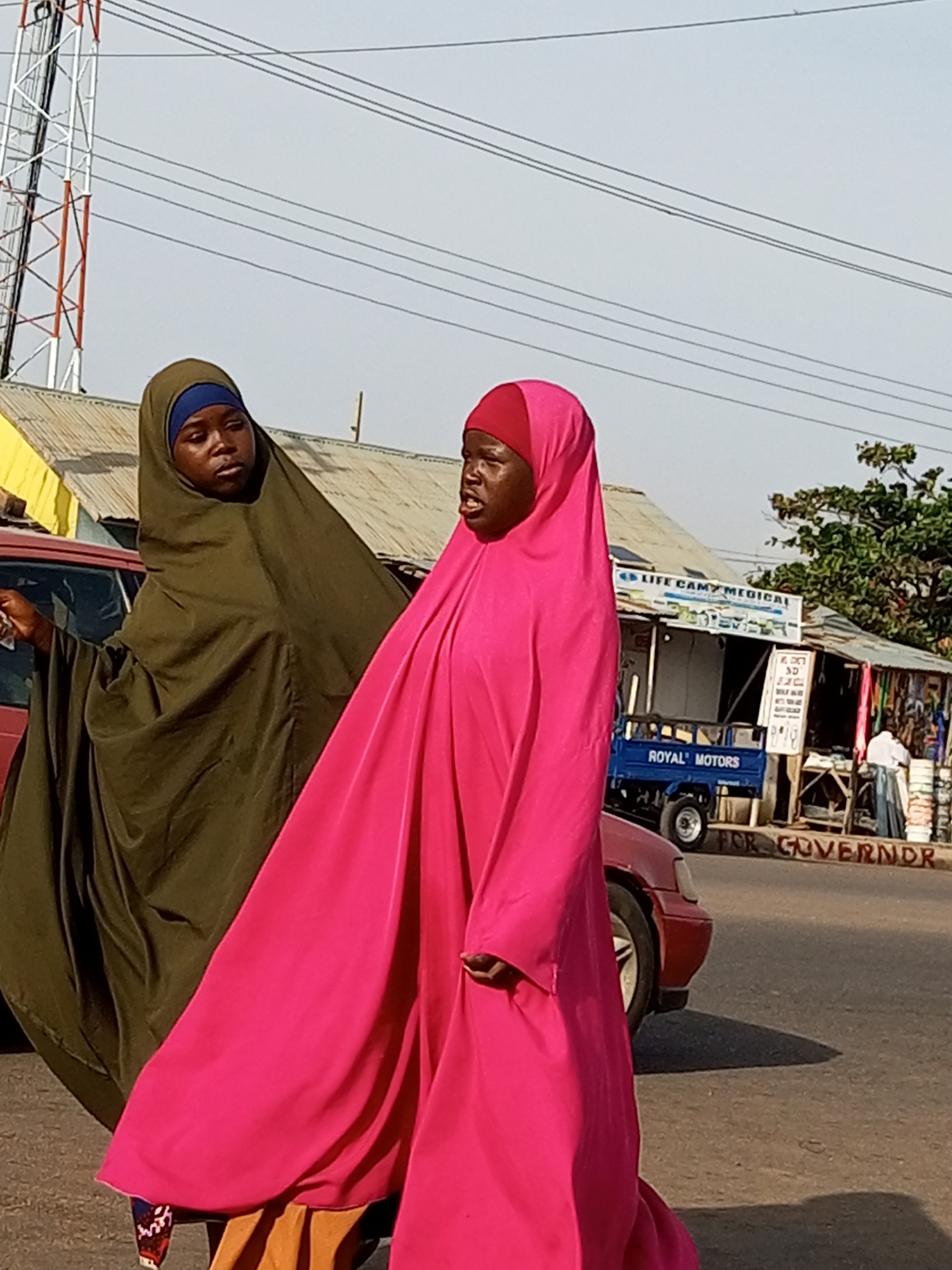 Two women wearing traditional Islamic head coverings and long, flowing dresses are walking on a street in Tempure, Nigeria. The woman on the left wears a dark olive green hijab and dress, while the woman on the right wears a bright pink hijab and dress. They are walking away from the camera, towards a row of buildings that appear to be shops. One building has a sign that reads "LIFE CAMP MEDICAL." A blue utility vehicle with "ROYAL MOTORS" written on its side is parked in front of one of the shops. There is a red car to the left of the women. In the background, a tall telecommunications tower is visible against a pale sky. The ground is paved and appears to be asphalt. Graffiti on the side of the road reads "FOR GOVERNR".