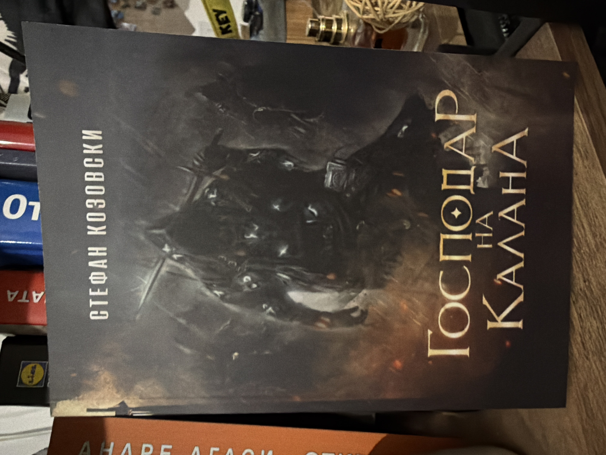 The image shows a book titled "Господар на Калана" (Lord of Kalana) by Стефан Козовски (Stefan Kozovski). The book cover features dark, abstract imagery. The book is situated among other books and objects, including a key and a decorative ball. The location appears to be indoors, possibly a home or library. There are no discernible details to specifically place this image in the city of Sofia, Bulgaria.