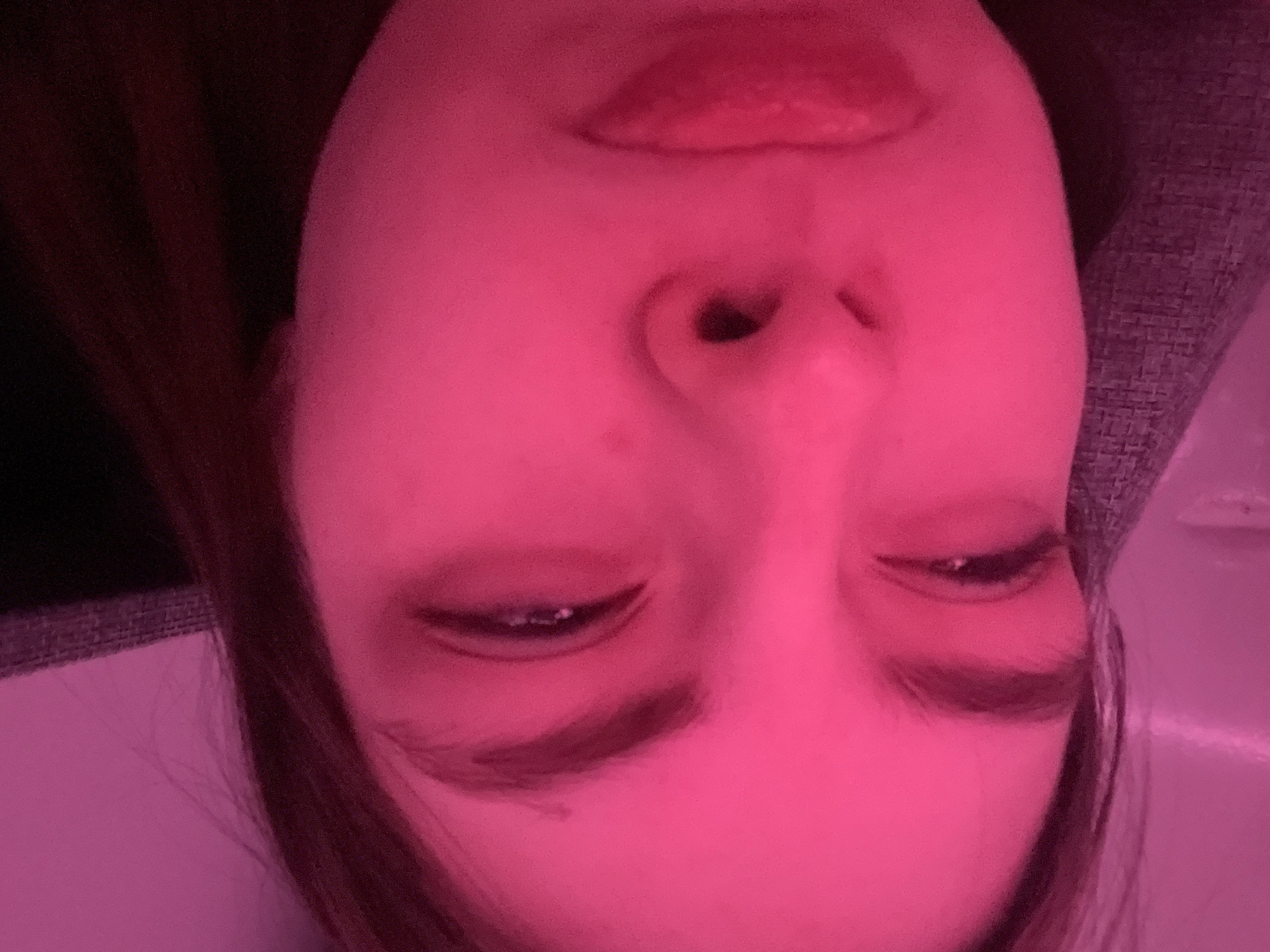 A close-up, upside-down view of a face is under a pink lighting. The subject's eyes are closed and their lips are slightly parted. The scene shows skin texture and features like eyebrows, eyelashes, and a nose. Background details are indistinct, with suggestions of hair and surfaces. There is no evidence in the image that suggests the location as Saint Petersburg, Russia, or any other specific location.