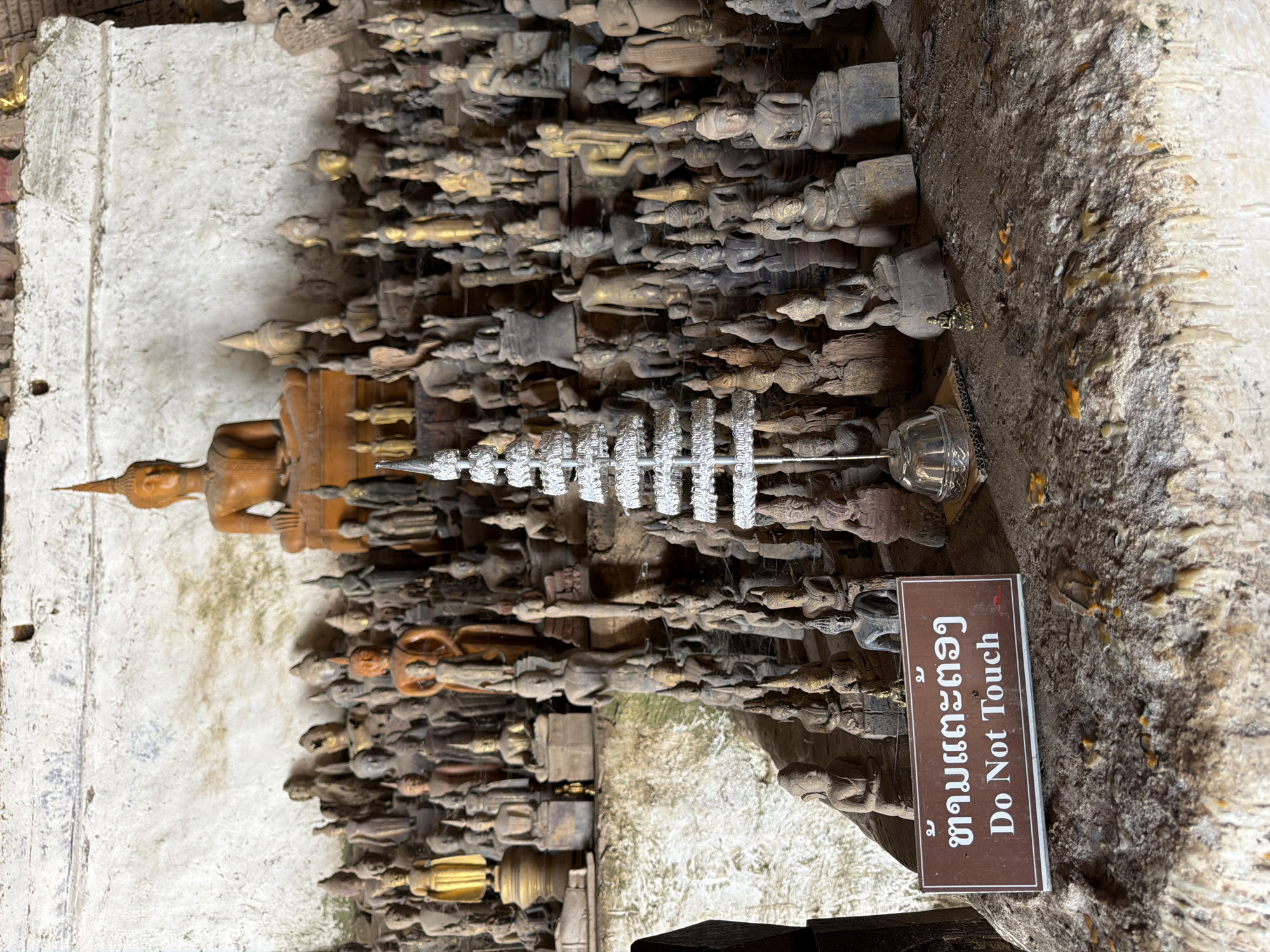 This image depicts a densely packed display of numerous small Buddha statues of various sizes, materials, and ages, situated within a cave or temple in Muangkeo, Laos. The statues are arranged on tiered surfaces and the ground, creating a crowded and layered scene. Many of the statues are made of wood or metal, with some appearing weathered and others more polished.
In the foreground, a brown sign with white text reads "Do Not Touch" in English, with Lao script above it. To the left of the sign, a small, intricate silver ornament resembling a tiered spire sits on a metal base. The environment appears to be a natural, earthy setting, with dirt and some organic matter visible on the ground. The wall behind the statues is rough and textured, with some discoloration and what might be moss or lichen growth in places. The lighting is dim, suggesting an indoor or shaded location. There are no people visible in the image. The overall impression is one of reverence and historical preservation, with the "Do Not Touch" sign indicating a need to protect these artifacts.