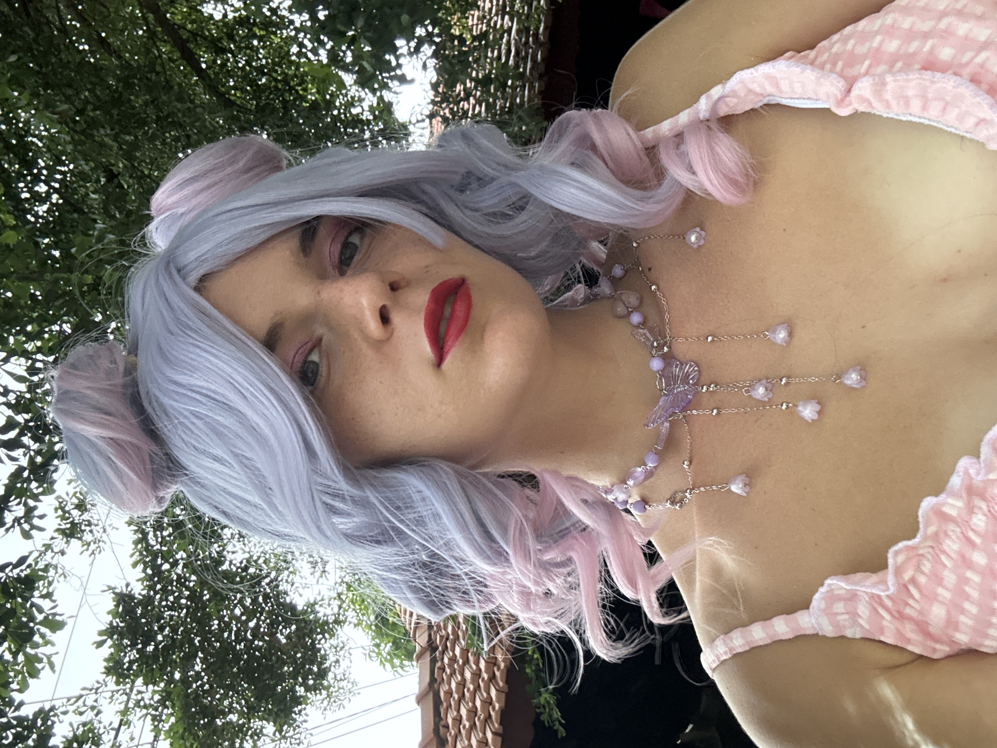 A young woman with pastel purple and pink hair styled into two buns and loose waves, wears a pink gingham top with ruffled straps and a delicate purple butterfly necklace with hanging floral charms. She has red lipstick and pink eyeshadow. She is outdoors, with dappled sunlight falling on her. Behind her, green foliage and a tiled roof are visible. The scene is likely in Mũi Né, Vietnam, a popular coastal resort town known for its beaches, sand dunes, and lush tropical vegetation.