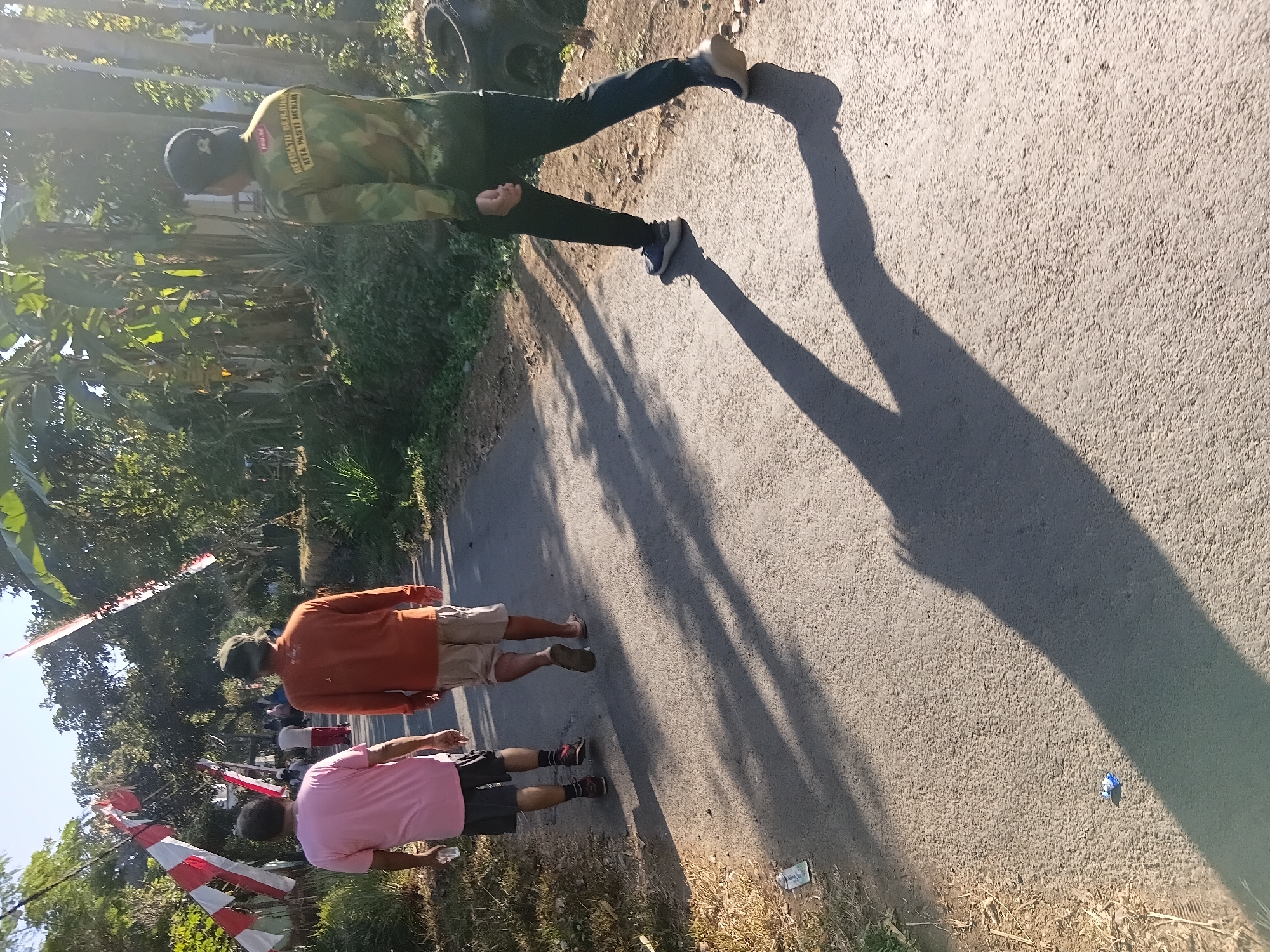 In Tanggul Wetan, Indonesia, three individuals walk along a paved road, casting long shadows in the late afternoon sun. The person on the left, wearing a pink t-shirt and dark shorts, carries a clear plastic bottle. To their right, another individual, in an orange long-sleeved shirt and light-colored shorts, walks with their back to the camera. A third person, dressed in a camouflage long-sleeved shirt and dark pants, walks further down the road in the same direction. The road is lined with trees and vegetation. Red and white flags, likely Indonesian flags, are visible on poles on the left side of the road. The scene appears to be a peaceful, everyday moment in a semi-rural setting.