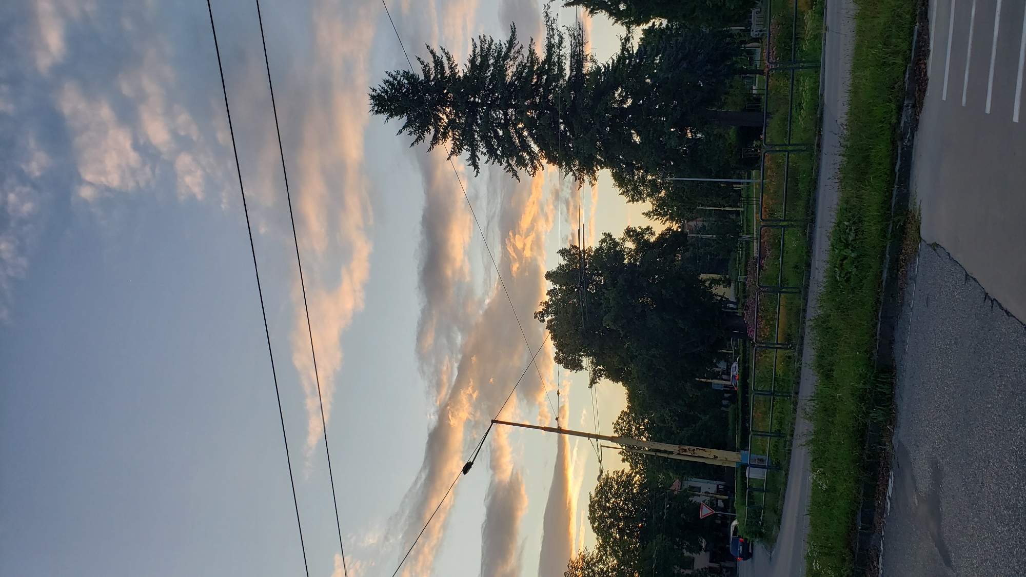 The image depicts a cloudy sky with hues of orange and pink, suggesting it was taken during sunset or sunrise. Power lines traverse the sky. In the foreground, a street curves to the left, with a grassy area and a metal fence separating it from a park-like setting. Several trees, including a tall evergreen, are visible. A lamppost stands near the street. In the background, buildings and more trees are present. The scene appears to be an urban environment in Prešov, Slovakia, possibly near a public space or intersection. No people or specific actions are discernible.