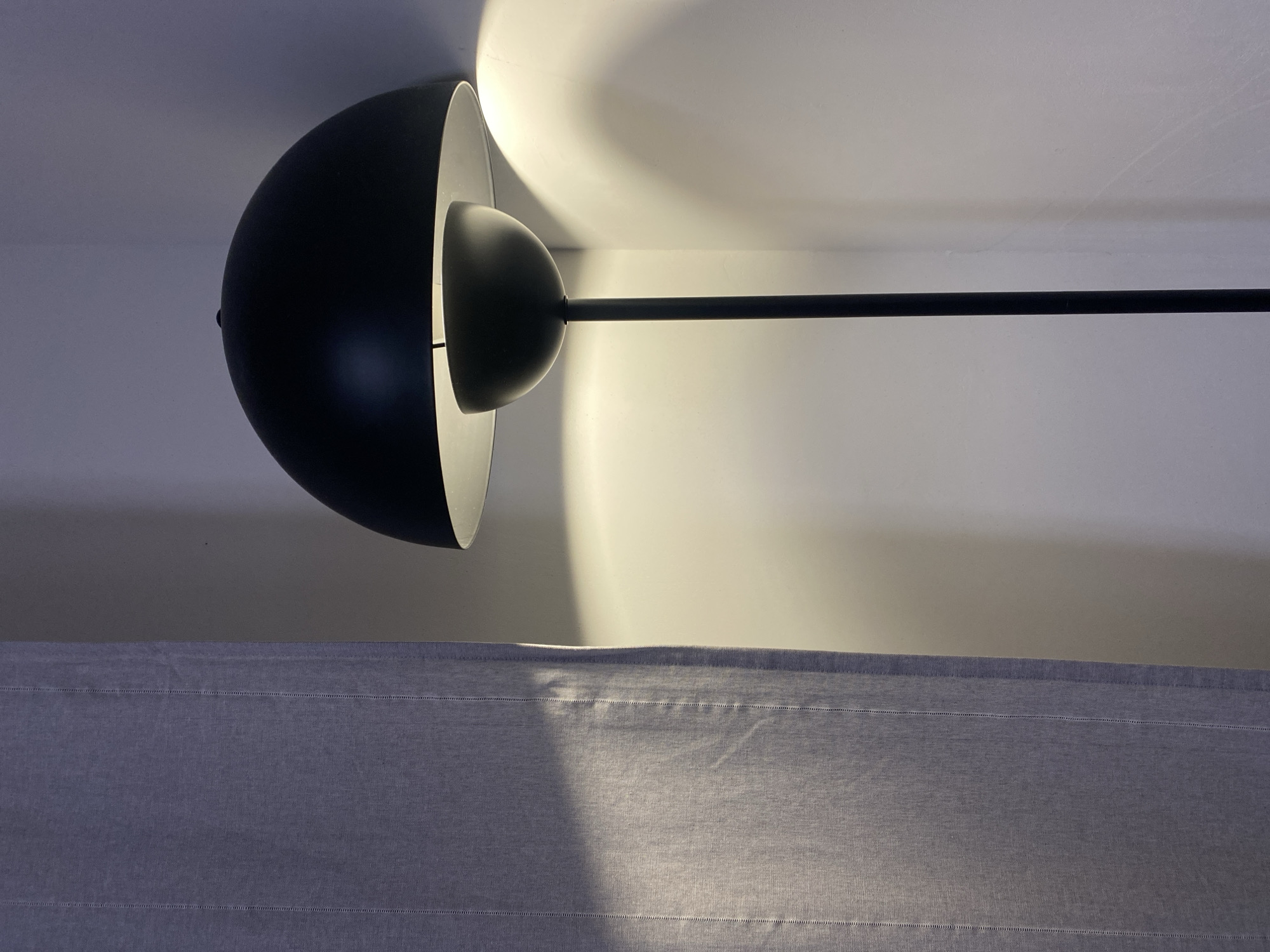 The image shows a black lamp with a spherical shade, attached to a straight pole, standing in front of a textured, grey fabric panel. The lamp is illuminated, casting a bright glow on the wall behind it. The scene appears to be indoors. The photograph is rotated 90 degrees clockwise. There is no evidence in the image to suggest it was taken in Izhevsk, Russia.