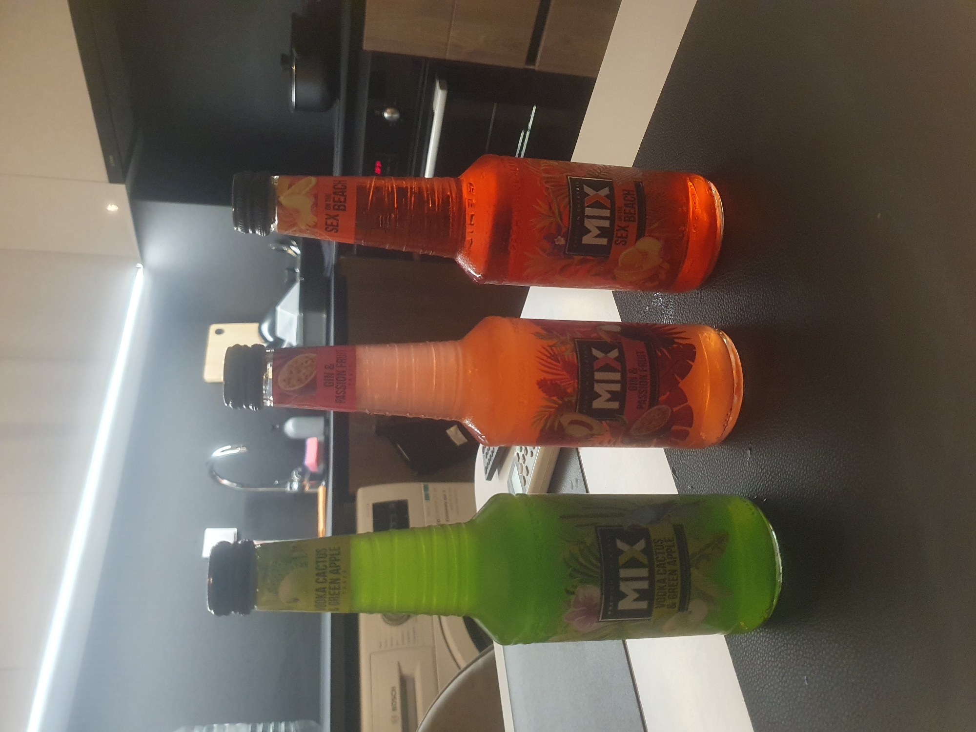 The image shows three bottles of "MIX" brand cocktails, positioned horizontally on a table with a black, textured surface. The top bottle is orange and labeled "Sex Beach". The middle bottle is a lighter shade of orange and reads "Gin & Passion Fruit". The bottom bottle is green and is labeled "Vodka Cactus & Green Apple". The background includes a kitchen environment with a sink, appliances, and cabinets visible. The image provides no information about the location of the city of Plovdiv, Bulgaria.
