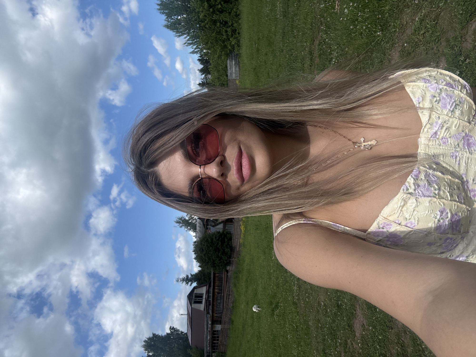 This is a portrait-oriented selfie taken outdoors. The primary subject is a young woman with long, blonde hair and pink lipstick. She is wearing red-tinted sunglasses and a light-colored dress with a floral pattern. Around her neck, she has a delicate necklace with a cross pendant.
The setting appears to be a rural area with a grassy field and trees in the background. There are also some wooden structures that resemble buildings or fences. The sky is blue with scattered white clouds, suggesting it is daytime and the weather is pleasant. The image is well-lit, indicating it was taken on a sunny day. There is no visible text in the image. The woman's pose and facial expression suggest a casual and relaxed mood. The overall impression is one of a sunny day spent outdoors in a peaceful, rural environment.