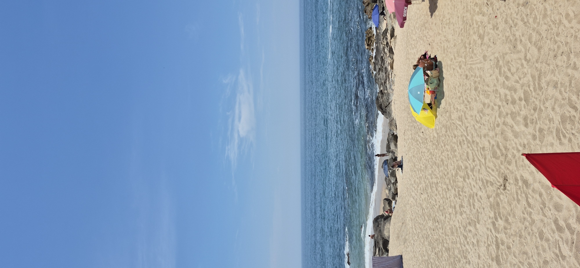 The image depicts a sunny day at Praia da Azurara beach in Vila do Conde, Portugal. The scene is divided into the sand, sea, and sky. The sand is light brown and textured, with footprints visible in some areas. Several beach umbrellas and tents are scattered across the sand, providing shade for beachgoers.
In the foreground, a bright red triangular object, possibly a flag or part of a beach shelter, is visible in the bottom left corner. A cluster of people is gathered under a yellow and blue umbrella towards the center of the image. One person is seated while others are lying down. A pink umbrella with the word "TENCE" is visible on the right side of the image.
Further back, closer to the water, more people are seen on the sandy shore and among the rocks. Some individuals are standing, while others are sitting or sunbathing. The water is a clear blue, with small waves breaking on the rocky shoreline. Large, dark rocks are scattered along the water's edge, interspersed with patches of sand. A striped beach shelter is positioned on the far left of the image. The sky is a clear, vibrant blue with a few wispy white clouds. The horizon line is sharp, separating the sea from the sky. The overall atmosphere is one of a relaxed summer day at the beach.