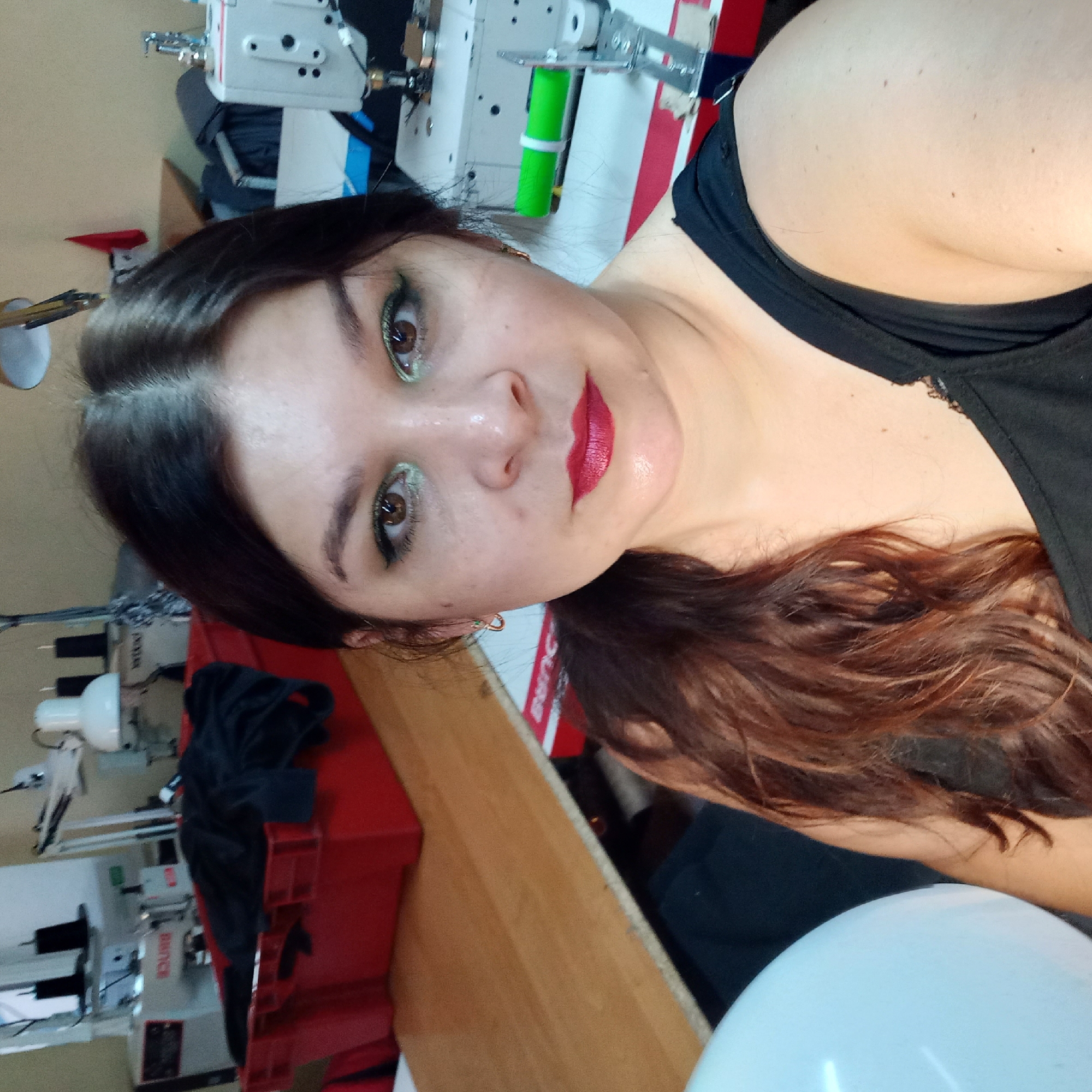 A medium shot photo shows a woman with dark brown hair, wearing a black tank top, and full makeup. She is sitting in what appears to be a sewing or tailoring workshop. The background features sewing machines, spools of thread, and a red plastic bin filled with dark fabric. A large white balloon is visible on the left side of the image. The lighting suggests an indoor setting. There is no discernible information within the image to indicate the location in Kropyvnytskyi, Ukraine.
