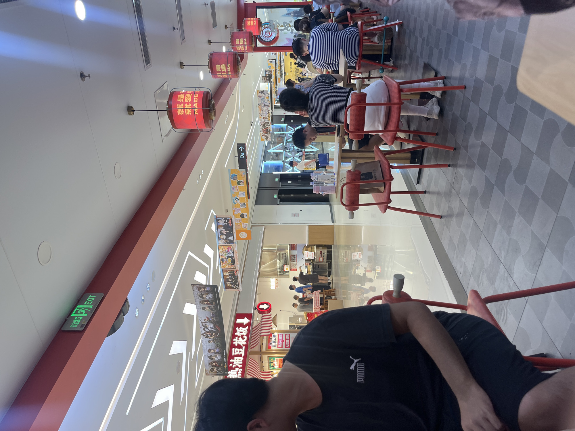 The image depicts a brightly lit, modern indoor dining area, likely a food court or a section of a shopping mall, situated in Yuzhong District, China. The scene is dominated by a row of diners seated at light wooden tables with distinctive red metal chairs.
The floor is covered in alternating light and dark grey square tiles arranged in a wave-like or interlocking pattern, exhibiting high reflectivity that mirrors overhead lights and subtle reflections of individuals. The ceiling is white, featuring a grid of bright linear lights, recessed circular lights, and air conditioning vents. A prominent red horizontal beam stretches across the upper part of the image, holding a green "EXIT" sign (出ロ) in both English and Chinese characters.
Overhead, several red cylindrical lanterns, adorned with Chinese characters (e.g., "就是东北爱" – "It's Northeast Love") and "MADE IN CHINA," hang from the ceiling, providing warm accent lighting. Decorative banners featuring anime or game characters are suspended from the ceiling structure throughout the space.
In the mid-ground, a food stall with a red and white striped awning displays a large red sign reading "热油豆花饭" (Hot Oil Tofu Rice) with a downward arrow. Smaller Chinese characters below translate to "Born for Chongqing, if you are not satisfied with the dish, you can return/exchange it without reason," explicitly identifying a local Chongqing specialty. Several individuals, likely staff, are visible behind the counter of this and other distant food stalls. Further into the background, a wide, brightly lit mall corridor with multiple entrances/storefronts and a "D→" directional sign can be seen, with pedestrians walking through.
In the immediate foreground to the left, a male individual is seated with his back partially to the camera, wearing a black Puma T-shirt and black shorts. His right arm rests on the table. Other diners in the mid-ground are casually dressed, seated at tables, and appear to be eating or conversing. One individual wears a black and white striped shirt. A brown paper bag hangs from the back of one of the red chairs. The overall environment is bustling, clean, and well-lit.