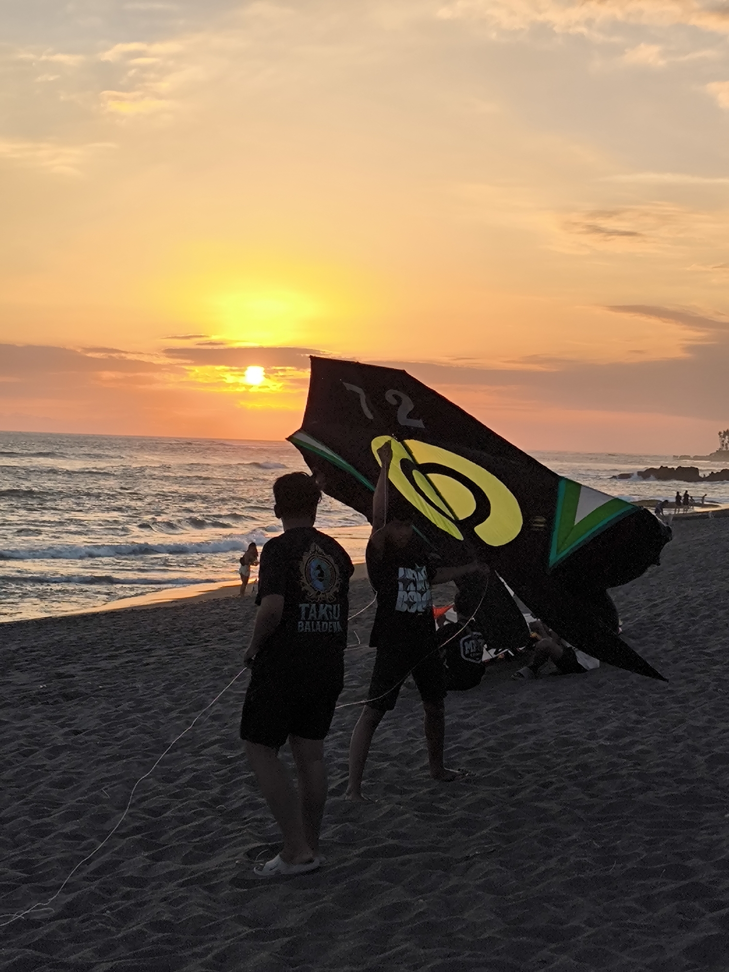 At dusk on a beach in Mengwi, Indonesia, two individuals are silhouetted against a vibrant orange and yellow sunset over the ocean. The person on the left, facing away from the camera, wears a black t-shirt with white lettering and black shorts. They hold a kite string that extends onto the dark sand. To the right, another person, also facing away, is in the process of launching or handling a large, black kite with yellow and green accents, including the number "72" and a circular symbol. The kite is partially obscuring the person to their left. The sandy beach is dark, with the texture of the sand clearly visible. Gentle waves break on the shore, and in the distance, a few other figures are visible along the water's edge, with some rocks and vegetation dotting the coastline further out. The sky is filled with soft, scattered clouds, painted with the warm hues of the setting sun.
