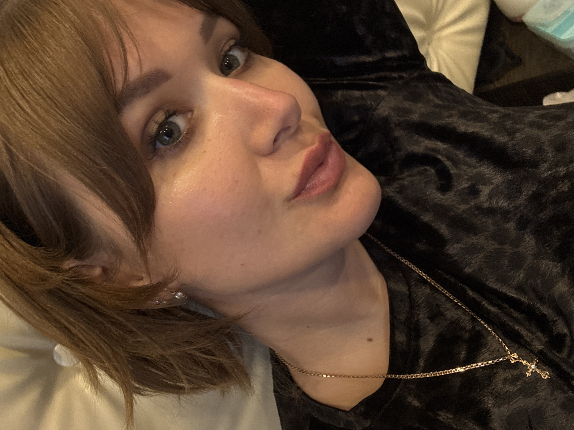A close-up portrait shows a young adult female, positioned with her head slightly tilted, gazing directly into the camera. Her medium-brown hair, styled with bangs, frames her face. She has light-colored eyes, possibly green or hazel, and full, reddish-pink lips with a subtle pout. Her skin appears clear with a natural sheen on her forehead and cheeks.
She is wearing a dark, possibly black, garment with a soft, velvet-like texture, suggesting an indoor setting like a home. A thin gold chain necklace with a small cross pendant, adorned with a tiny clear stone, rests on her chest. Small, sparkling earrings are visible on her left ear. The background is softly blurred, showing a light-colored pillow or cushion behind her head and darker, indistinct elements to her right, possibly furniture. The lighting suggests an indoor, possibly evening, environment. There is no visible text in the image. The overall impression is one of a relaxed pose for a portrait or selfie.