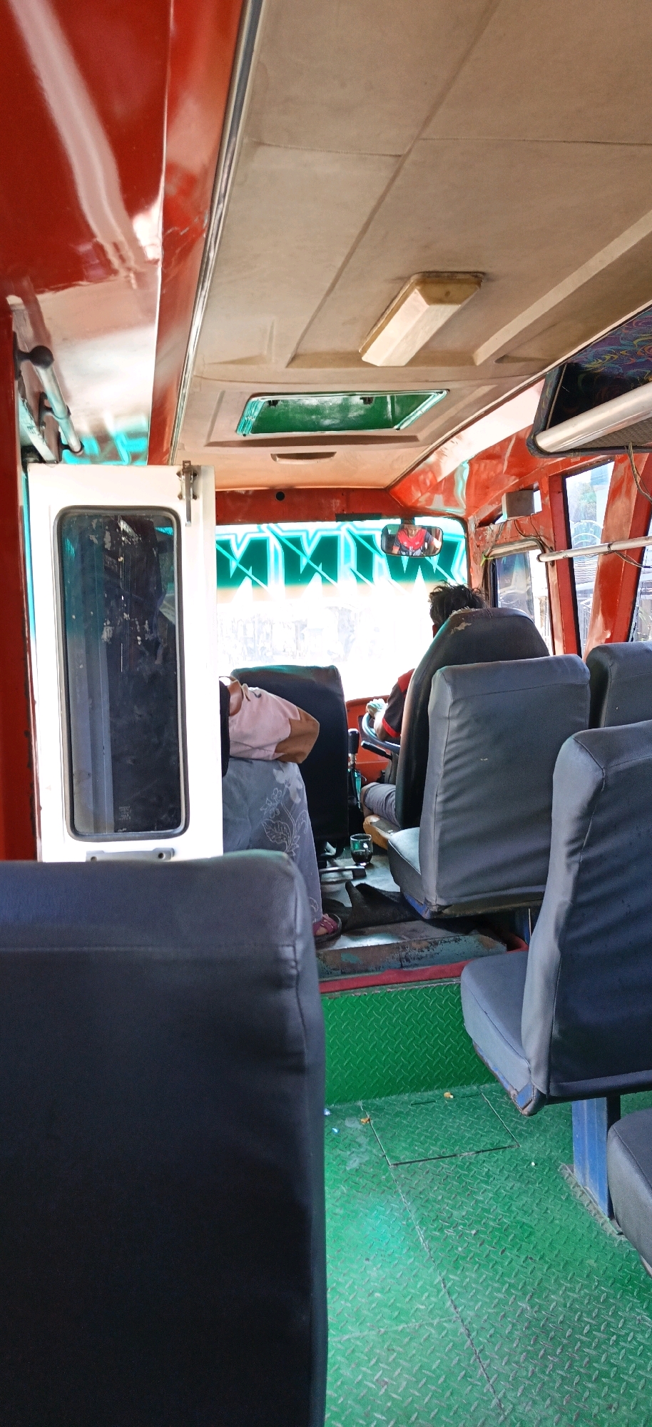 The image shows the interior of a bus, likely in Kebumen, Indonesia, viewed from the back seats looking forward. The bus has red trim and a beige ceiling with a light fixture. The floor is green diamond plate. There are several rows of dark gray seats. In the front, a driver is visible behind the steering wheel, wearing a red and black shirt and looking in a rearview mirror. To the left of the driver, a person in a pink shirt is seated and leaning back, with their legs crossed. A white door with a glass panel is open on the left side of the bus. Bright green lettering is visible on the front windshield.