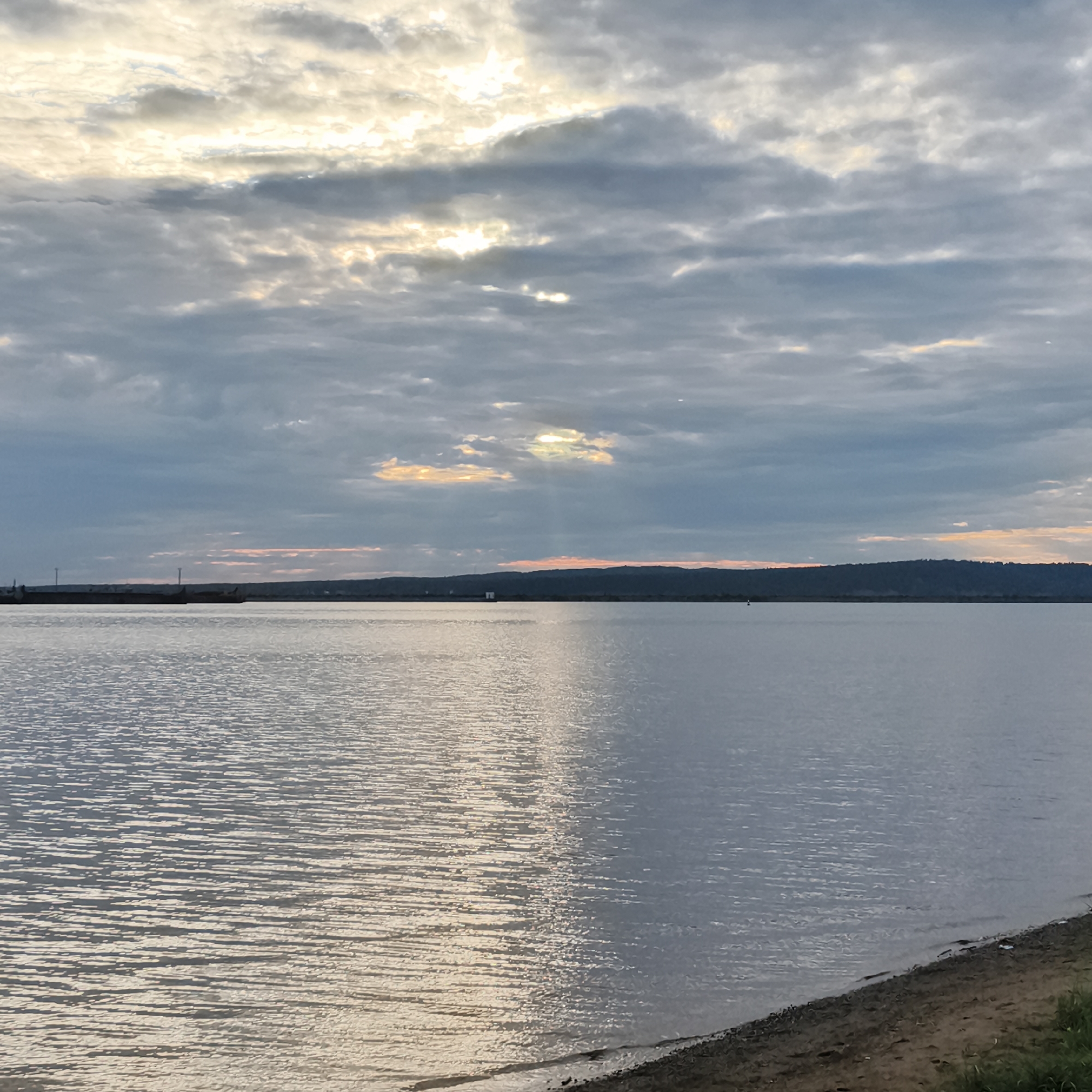 The image depicts a wide, calm body of water, likely a river or reservoir, with a shoreline in the foreground composed of sand and some sparse green vegetation. A silhouetted treeline or hill forms the horizon in the distance, with some structures, possibly industrial or navigational, visible along the shoreline. The sky is overcast with dramatic clouds, through which sunlight is breaking, creating rays of light and patches of warm color, suggesting either sunrise or sunset. The overall atmosphere is serene and natural. This scene is consistent with locations along the Kama River in Chaikovsky, Russia, known for its industrial port and scenic river views. There are no people or animals visible in the image.