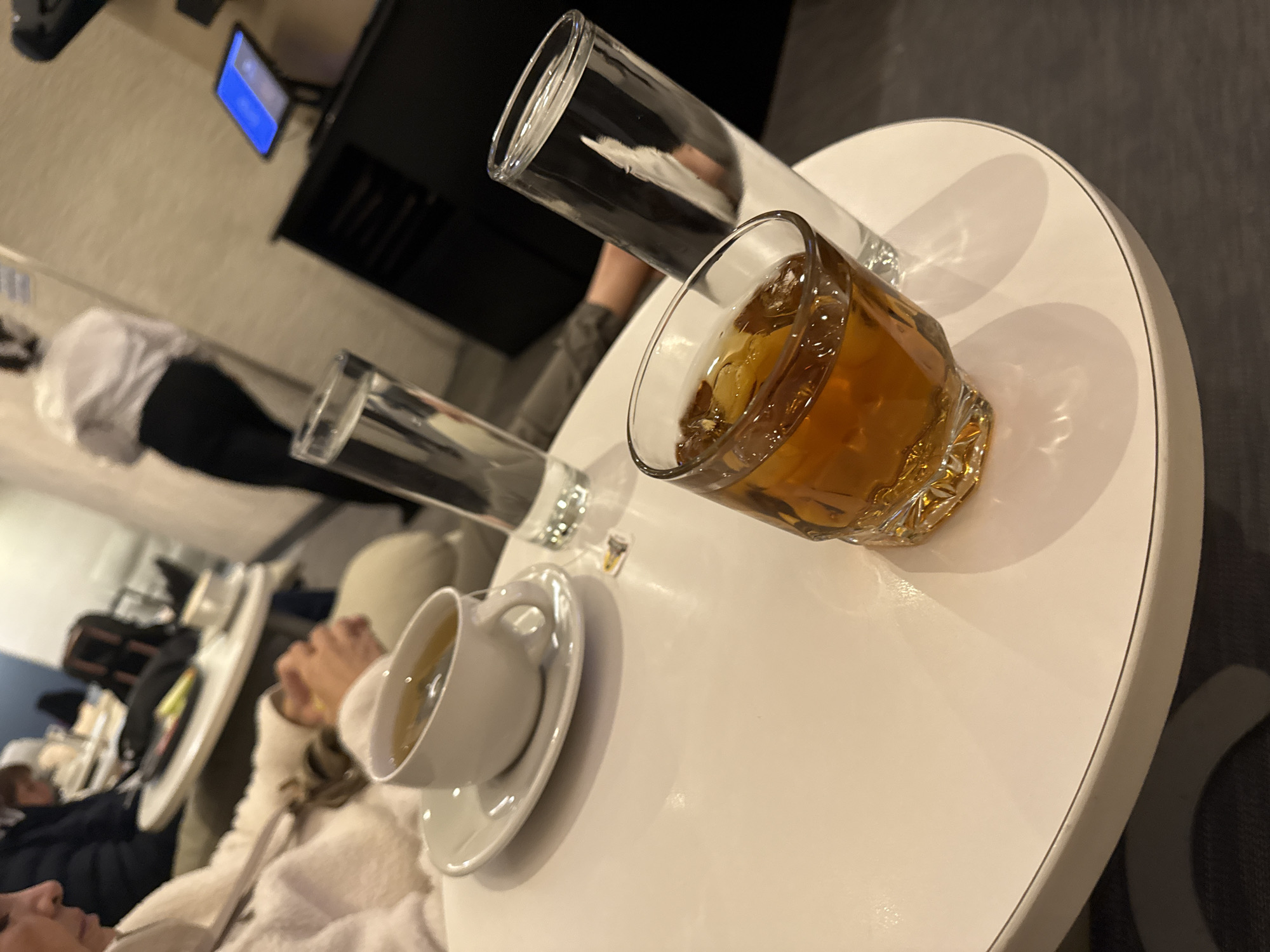 A round white table holds a cup of tea on a saucer, a glass of water, and a rocks glass of whiskey with ice. A tall, thin glass is behind the rocks glass. In the background are other people, a television, and a waiter serving. The location is likely an indoor seating area of a restaurant or lounge. The presence of a waiter and drinks suggests a hospitality venue. There is nothing that indicates the location is in Mexico City.